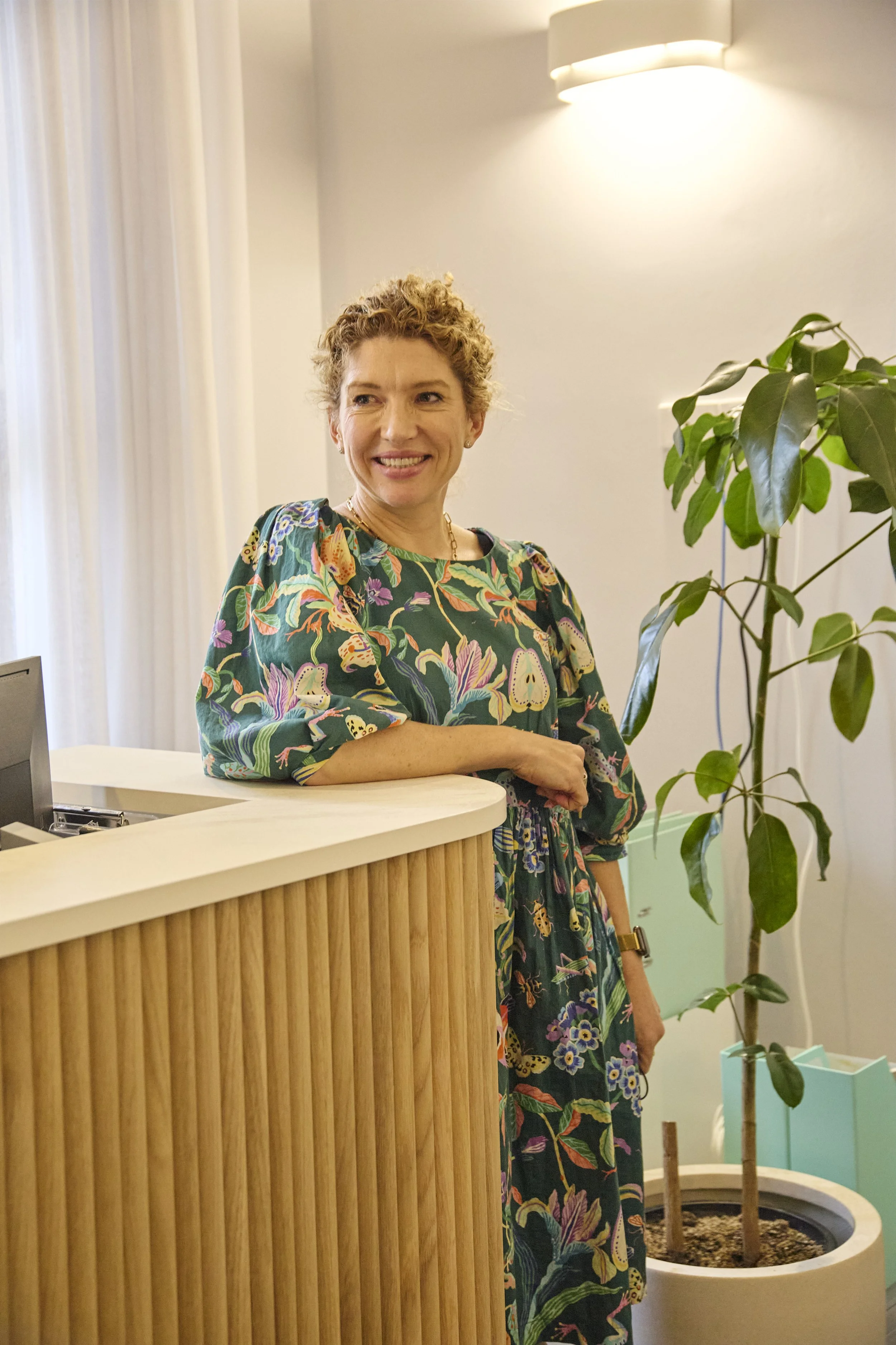 A woman with curly blonde hair smiling while standing behind a curved wooden reception desk, wearing a colorful floral dress, in an indoor space with white walls and a large green potted plant nearby.
