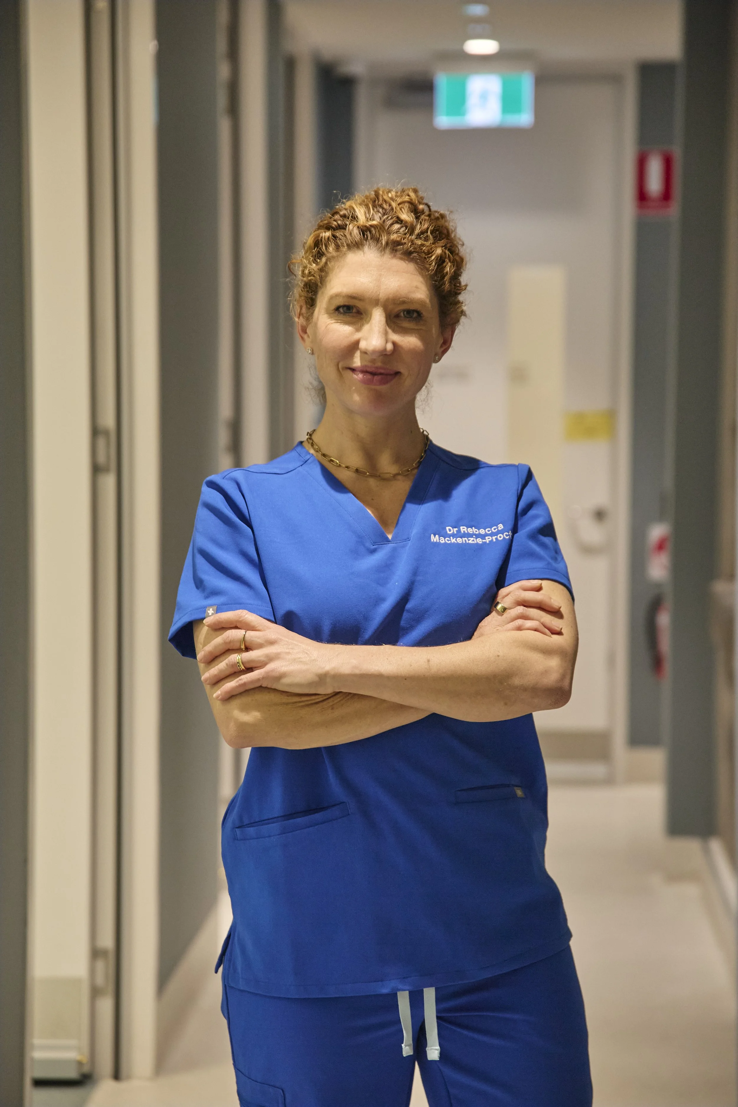 A woman with curly red hair wearing blue medical scrubs standing in a hospital corridor with arms crossed.