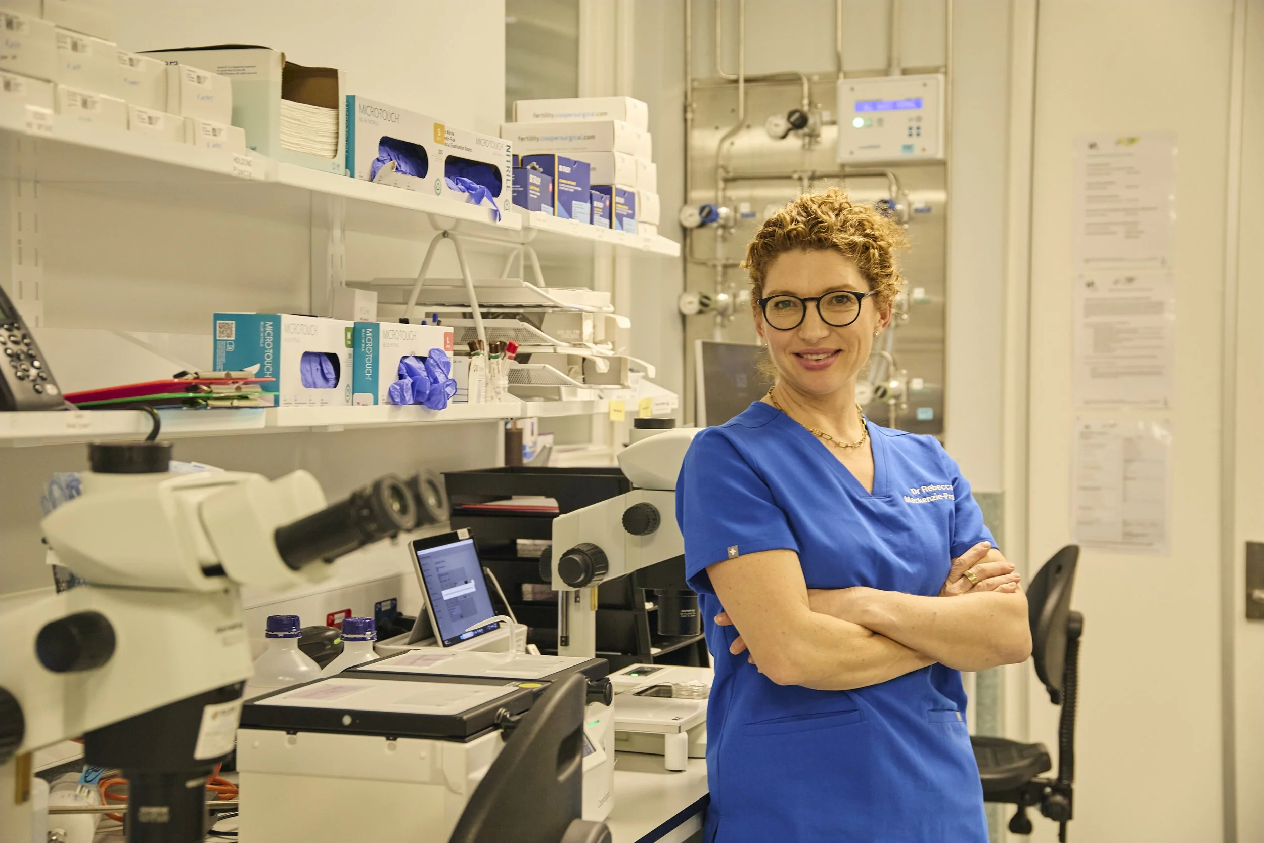 A woman in blue medical scrubs and glasses standing in a laboratory, smiling with arms crossed, surrounded by microscopes, laboratory equipment, and medical supplies.