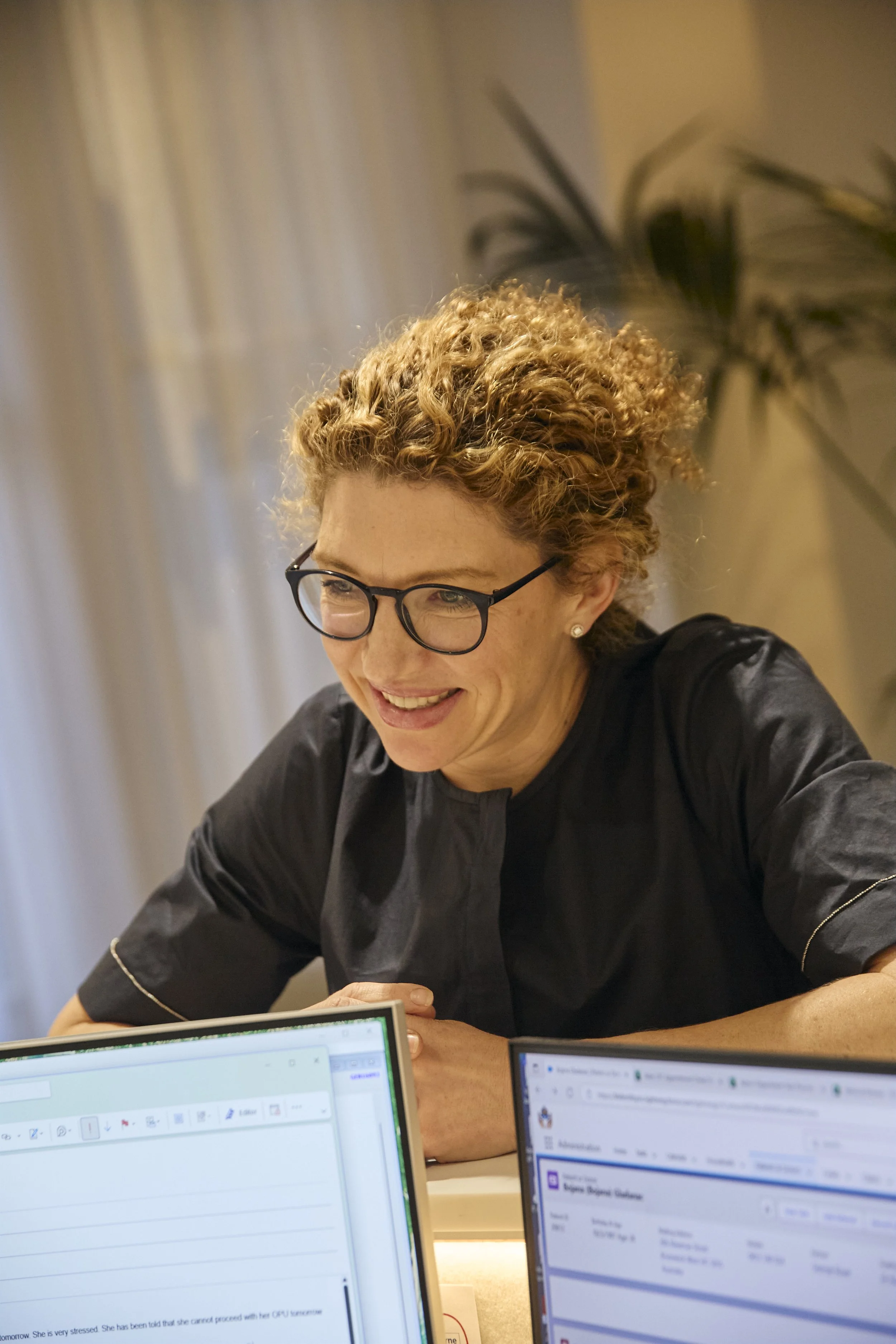 A woman with curly hair, glasses, and earrings smiling while looking at her laptop in an office setting with two open laptops in front of her.