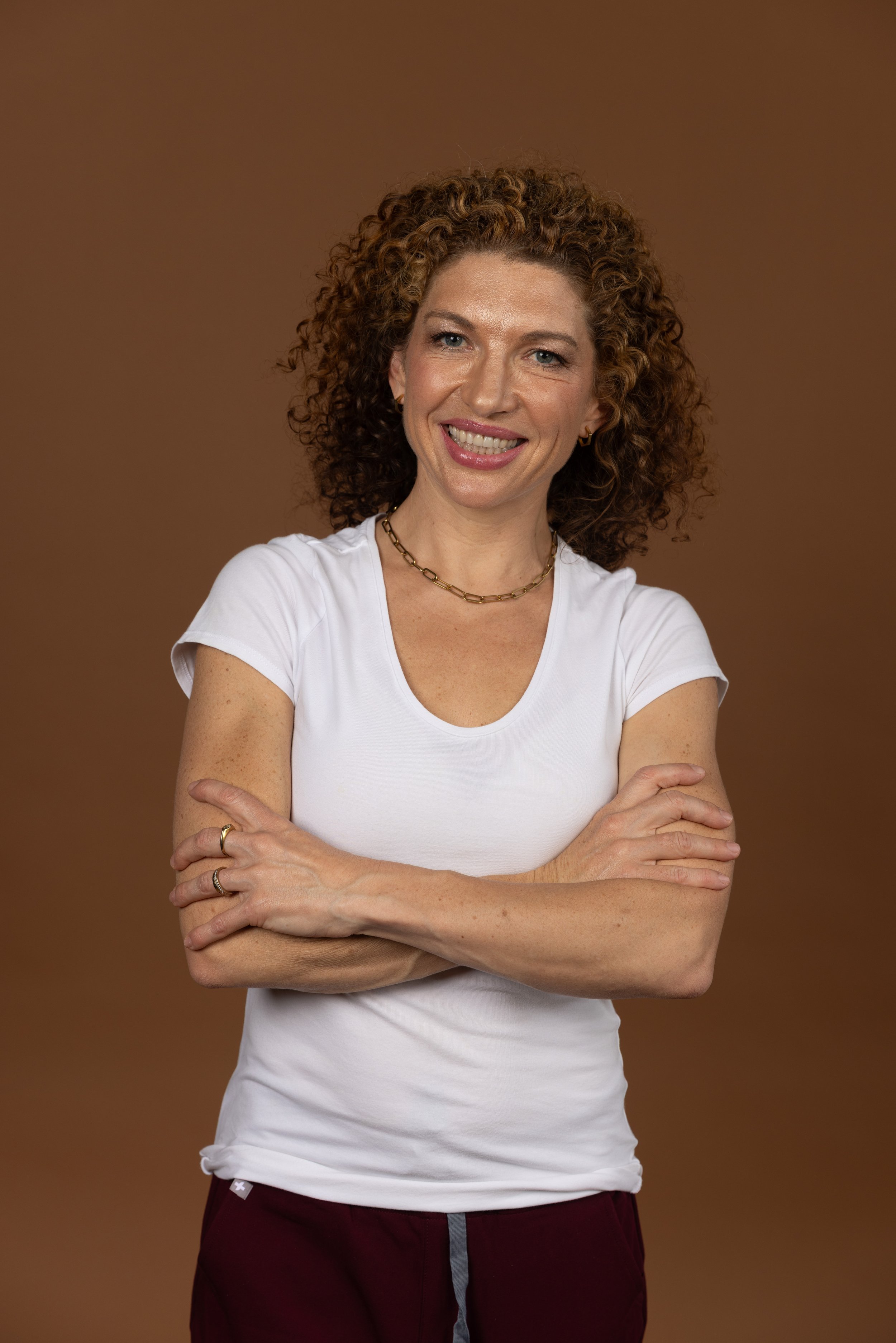 A woman with curly red hair smiling, wearing a white t-shirt and maroon pants, with a brown background.