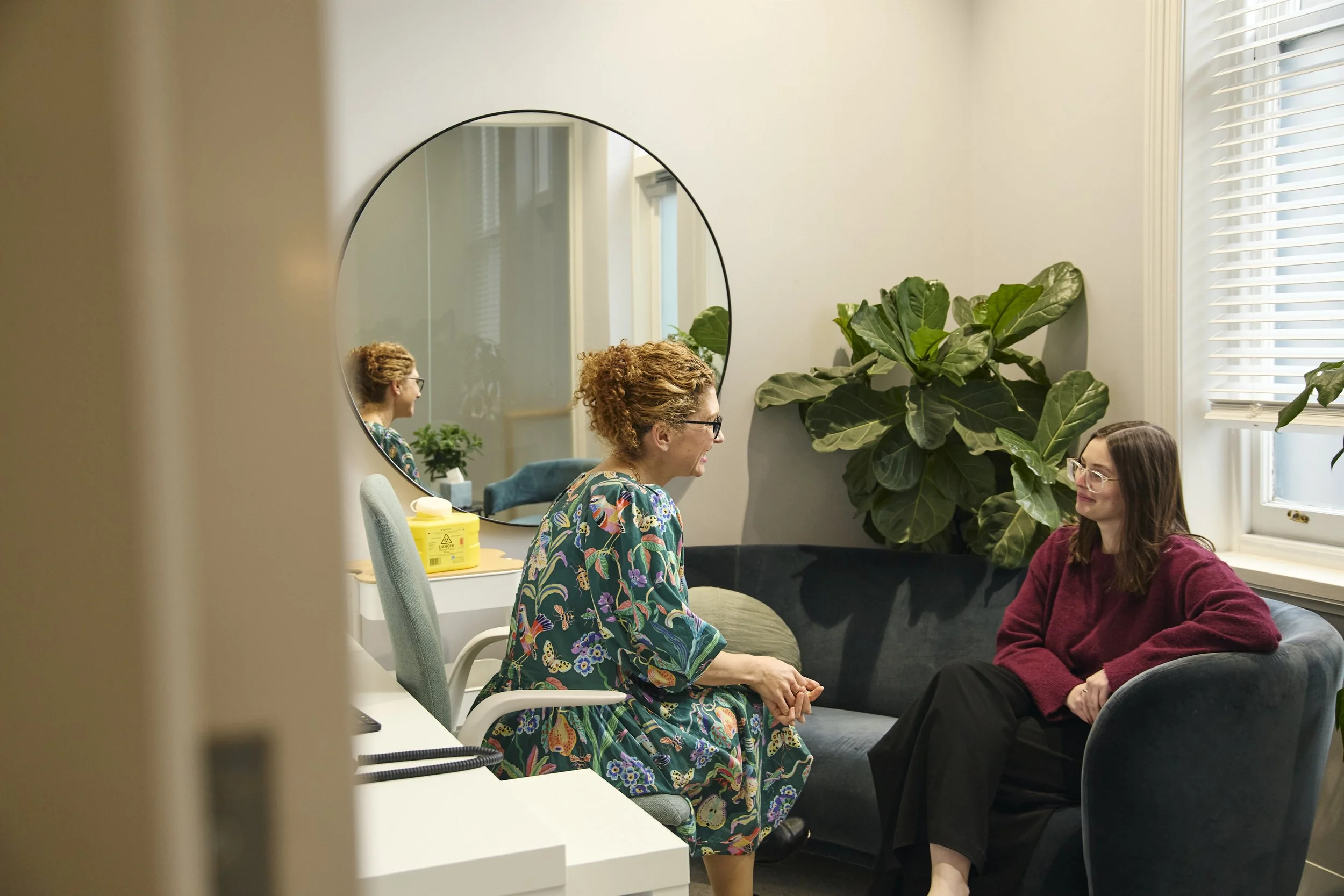 Two women having a conversation in an office, one sitting on a dark blue sofa and the other on an office chair, with a large leafy plant behind them and a round mirror on the wall.