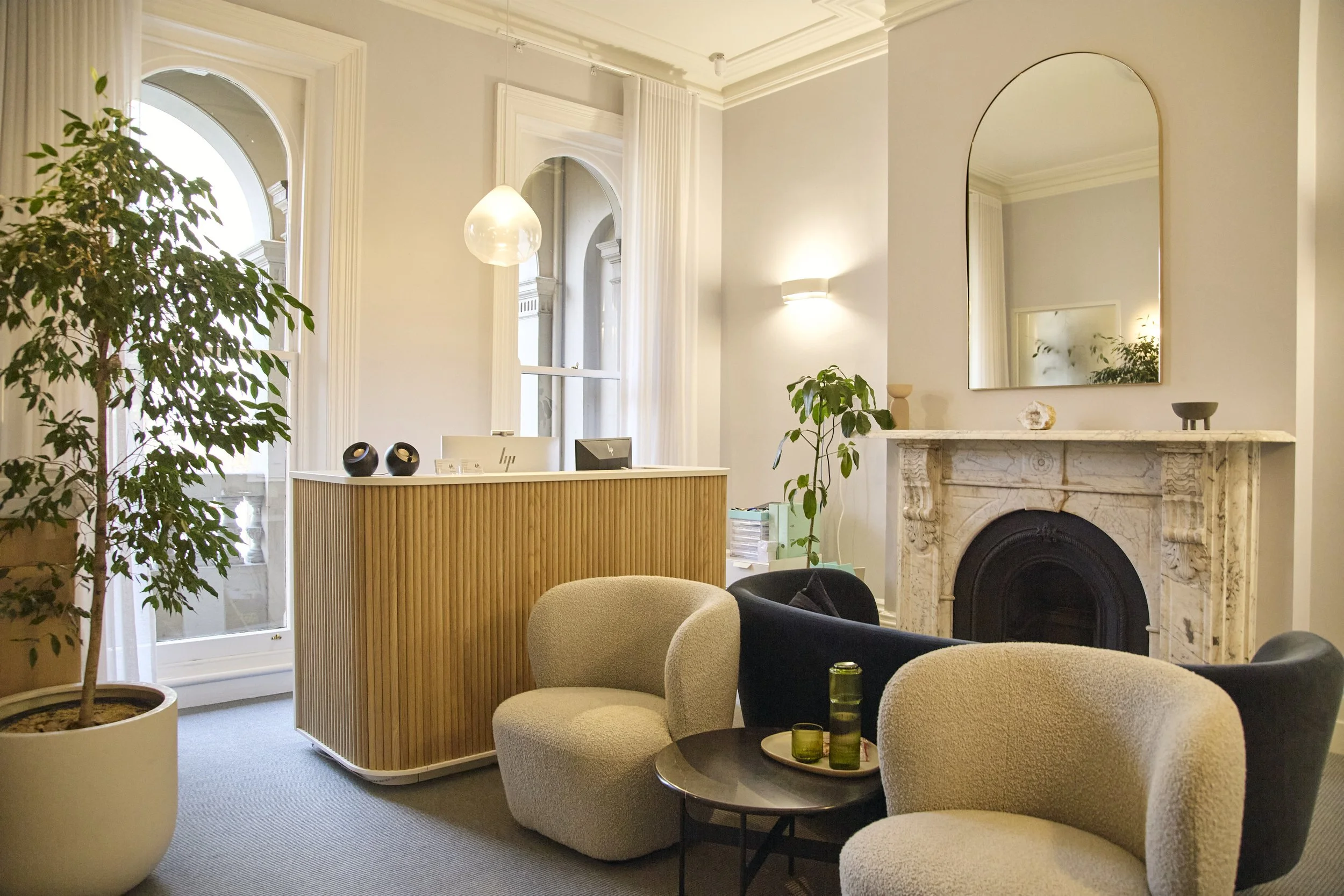 A cozy, stylish living room with large windows, a white fireplace with a marble mantel, a mirror above the fireplace, comfortable beige and black chairs around a small round table with green glasses, potted plants, and a reception desk with a computer, speakers, and files.