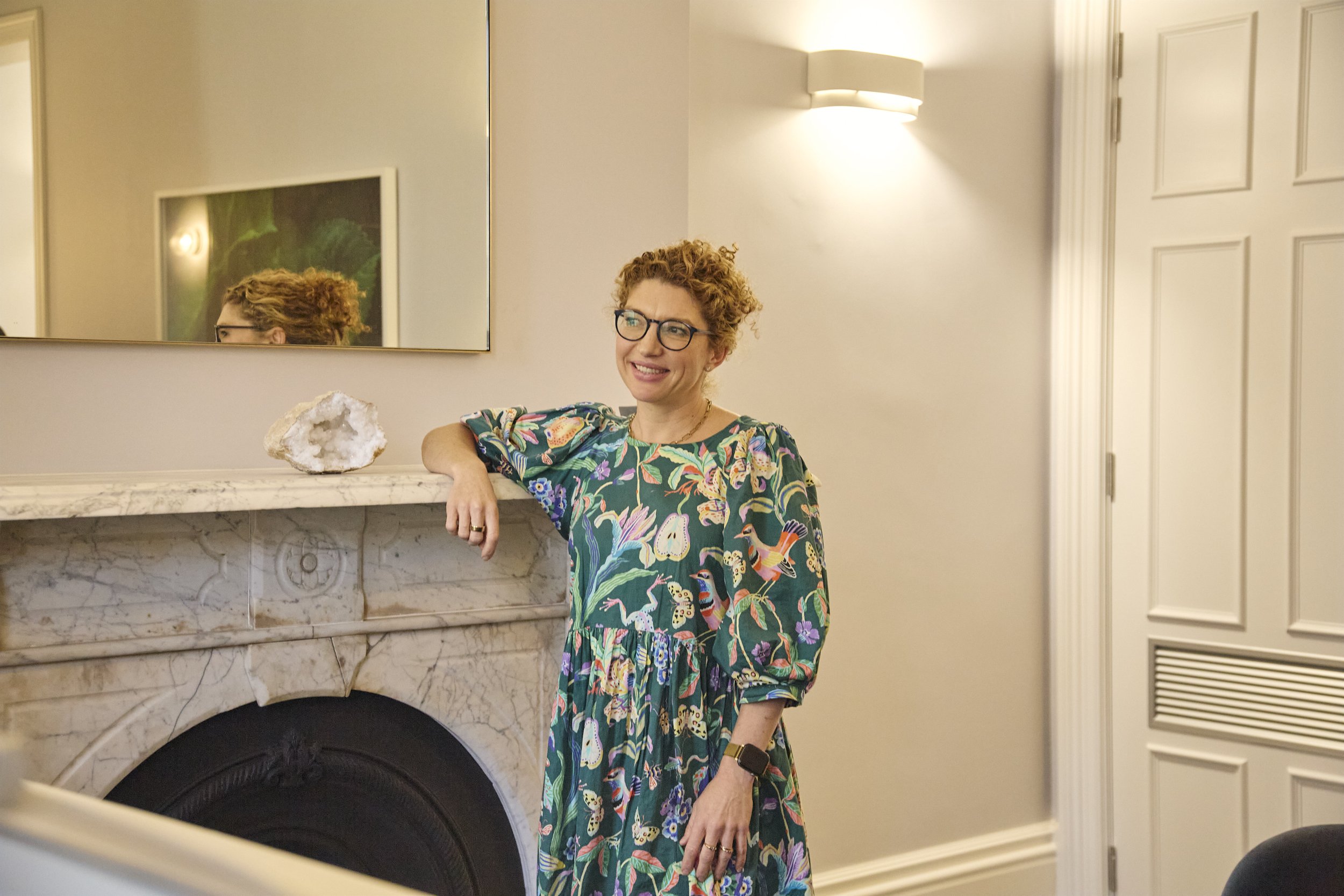 A woman with curly red hair and glasses standing by a fireplace with her arm resting on the mantelpiece, smiling, in a colorful floral dress in a well-lit room.