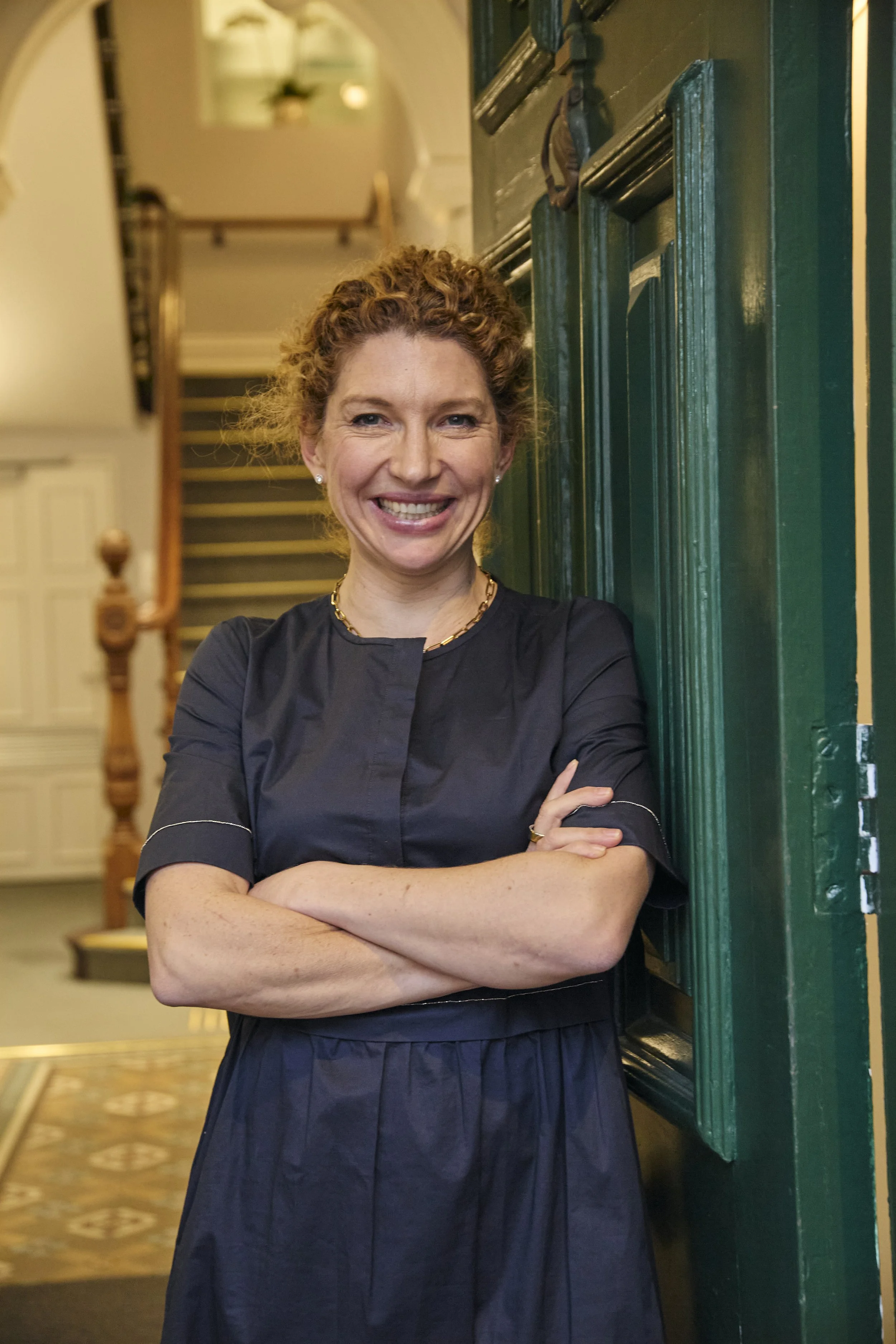 A woman with curly red hair smiling and standing with arms crossed next to a green door inside a house.