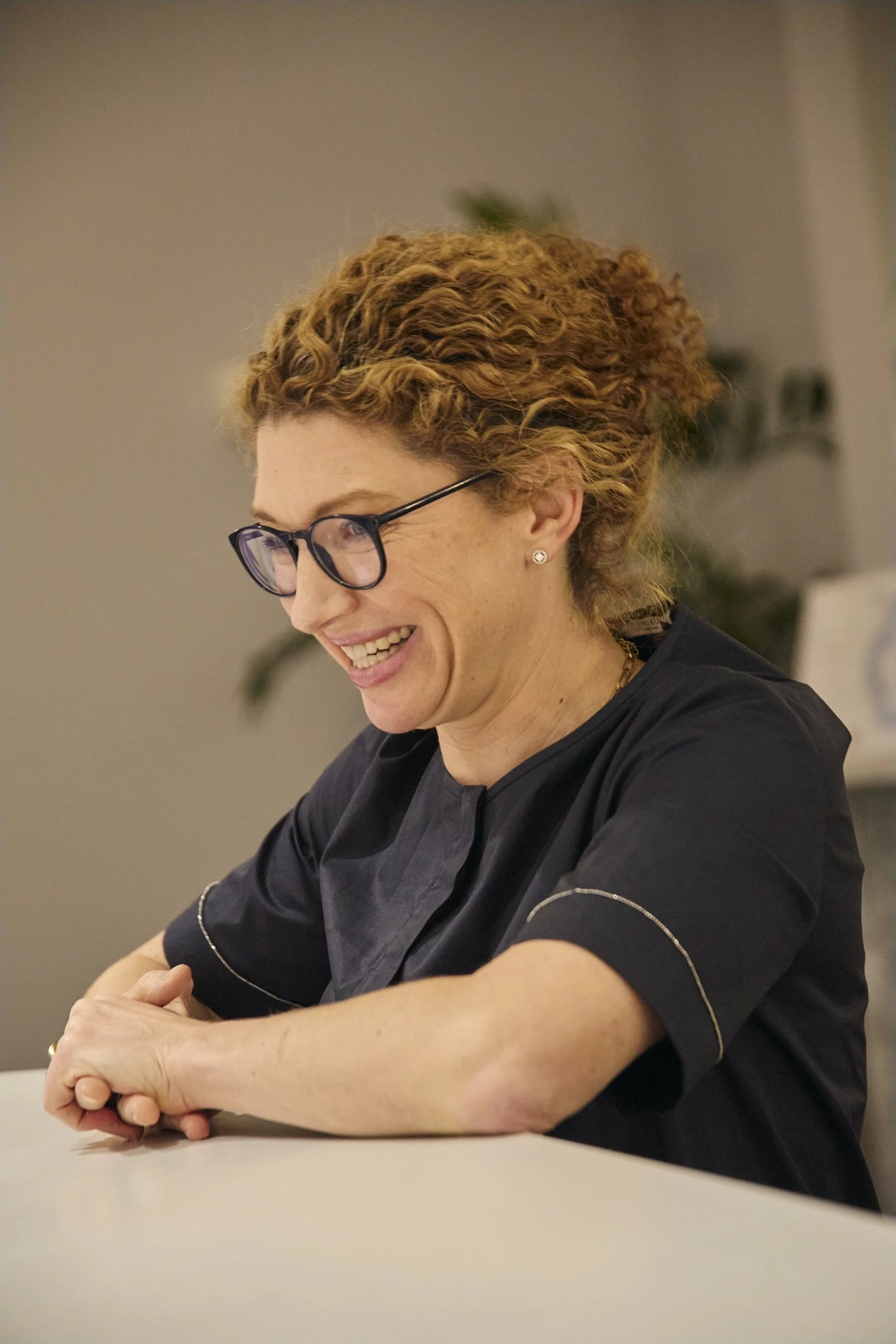 A woman with curly red hair wearing glasses and a dark professional uniform, smiling and leaning on a table.