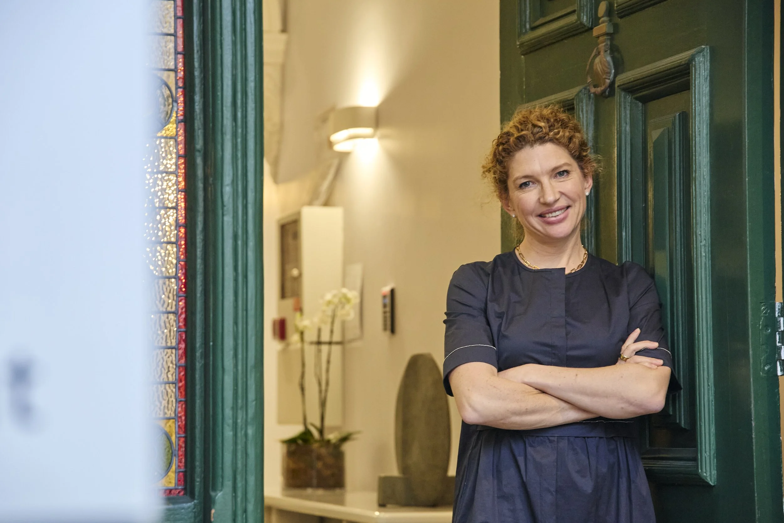 A woman with curly red hair smiling and standing with her arms crossed at a doorway with green trim.