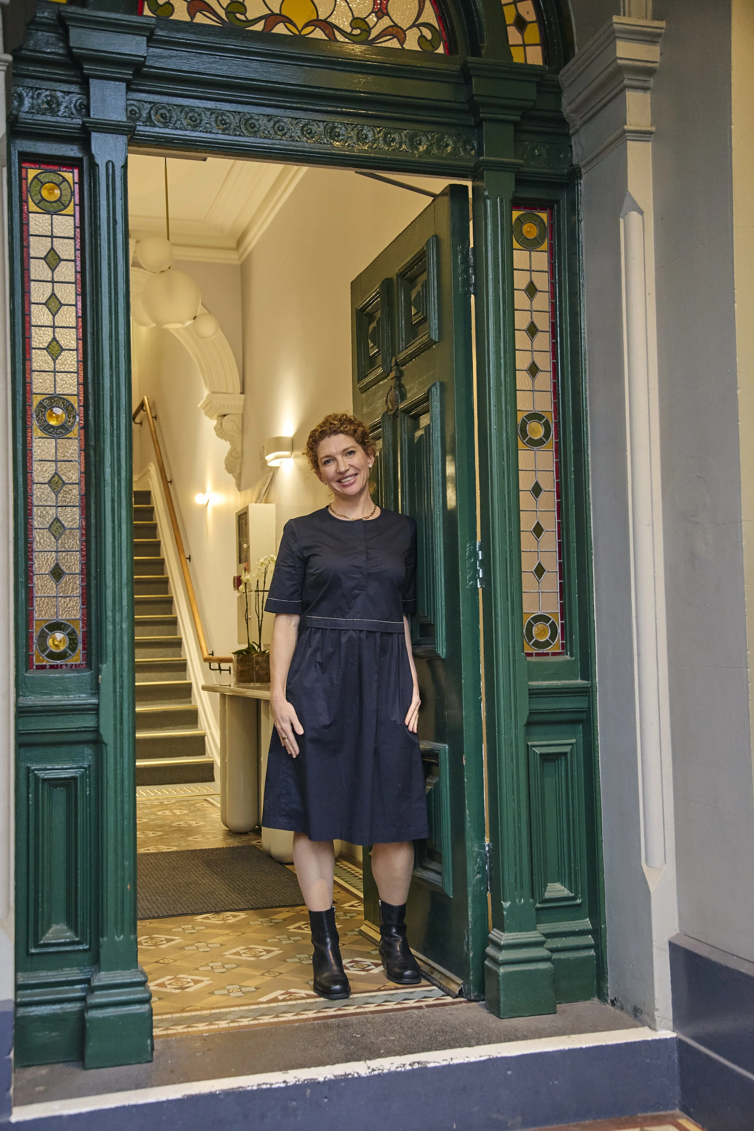 A woman standing at a doorway in a vintage-styled building, smiling at the camera. She has curly hair and wears a black dress with black boots. The door is green with stained glass panels.