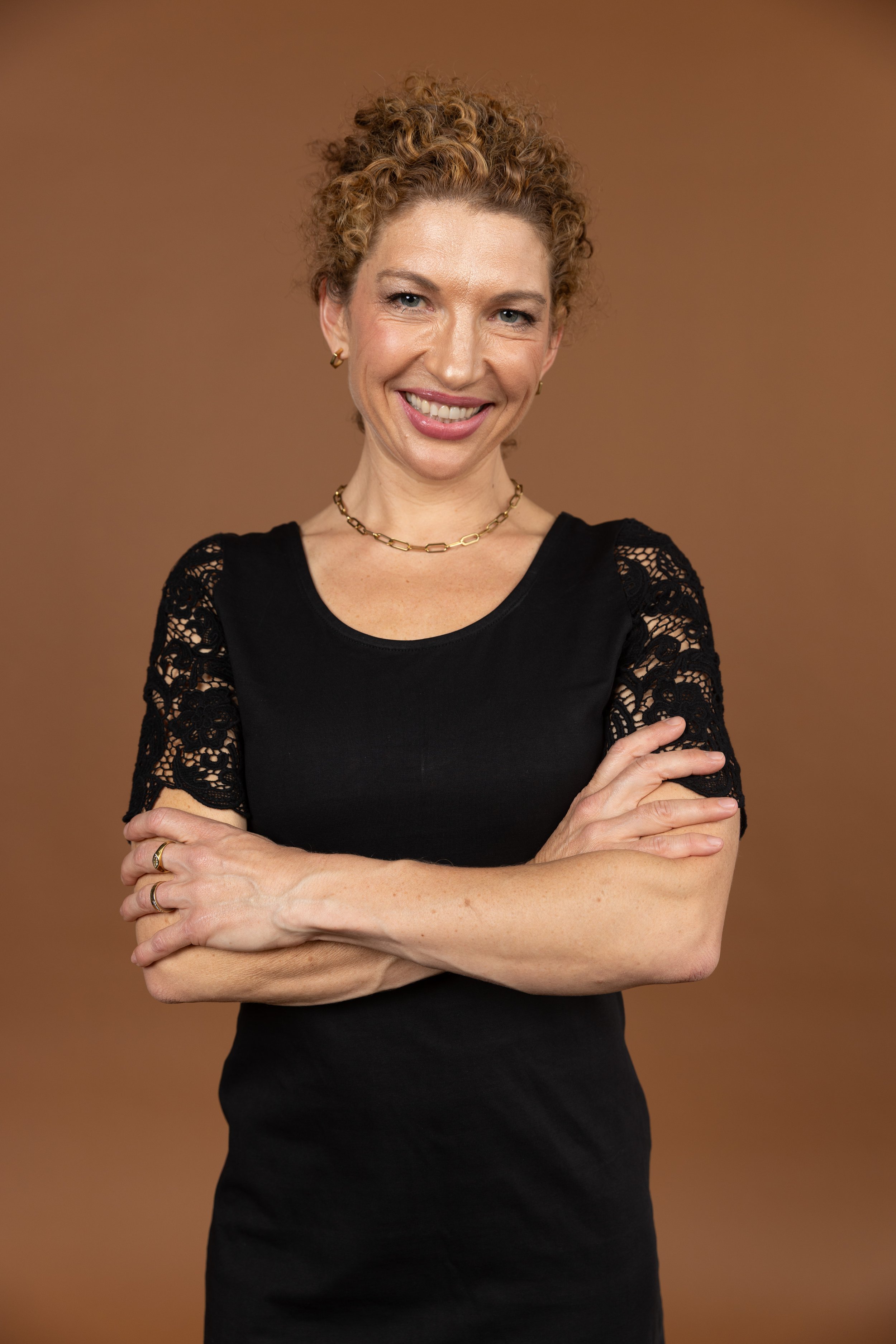 A woman with curly blonde hair, wearing a black dress with lace sleeves, gold jewelry, and smiling with crossed arms, standing against a brown background.