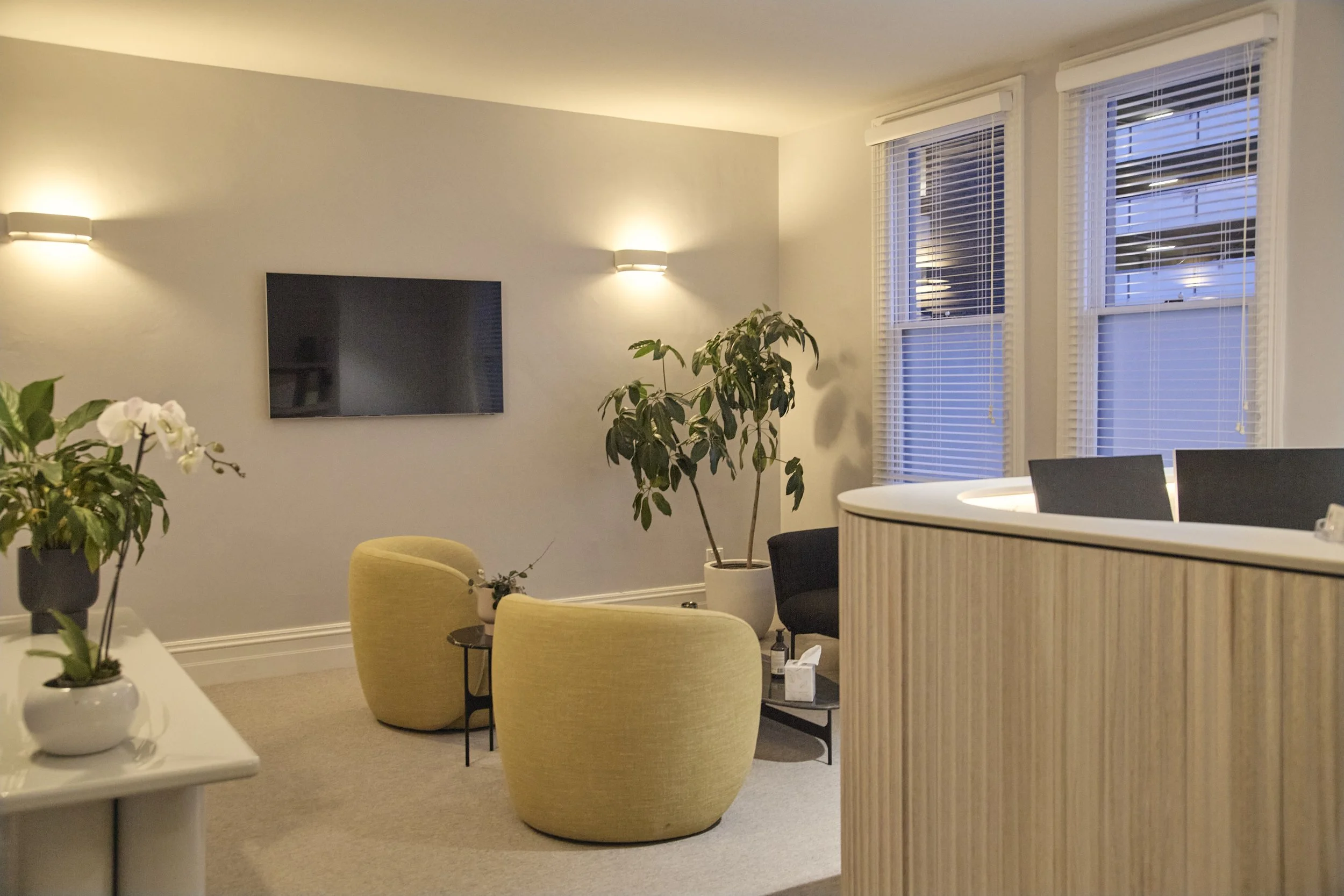 A modern living room with two yellow armchairs, a black side table, and a large potted plant near the windows. Two wall-mounted lights, a wall-mounted TV, and a white counter with two black chairs are also visible.