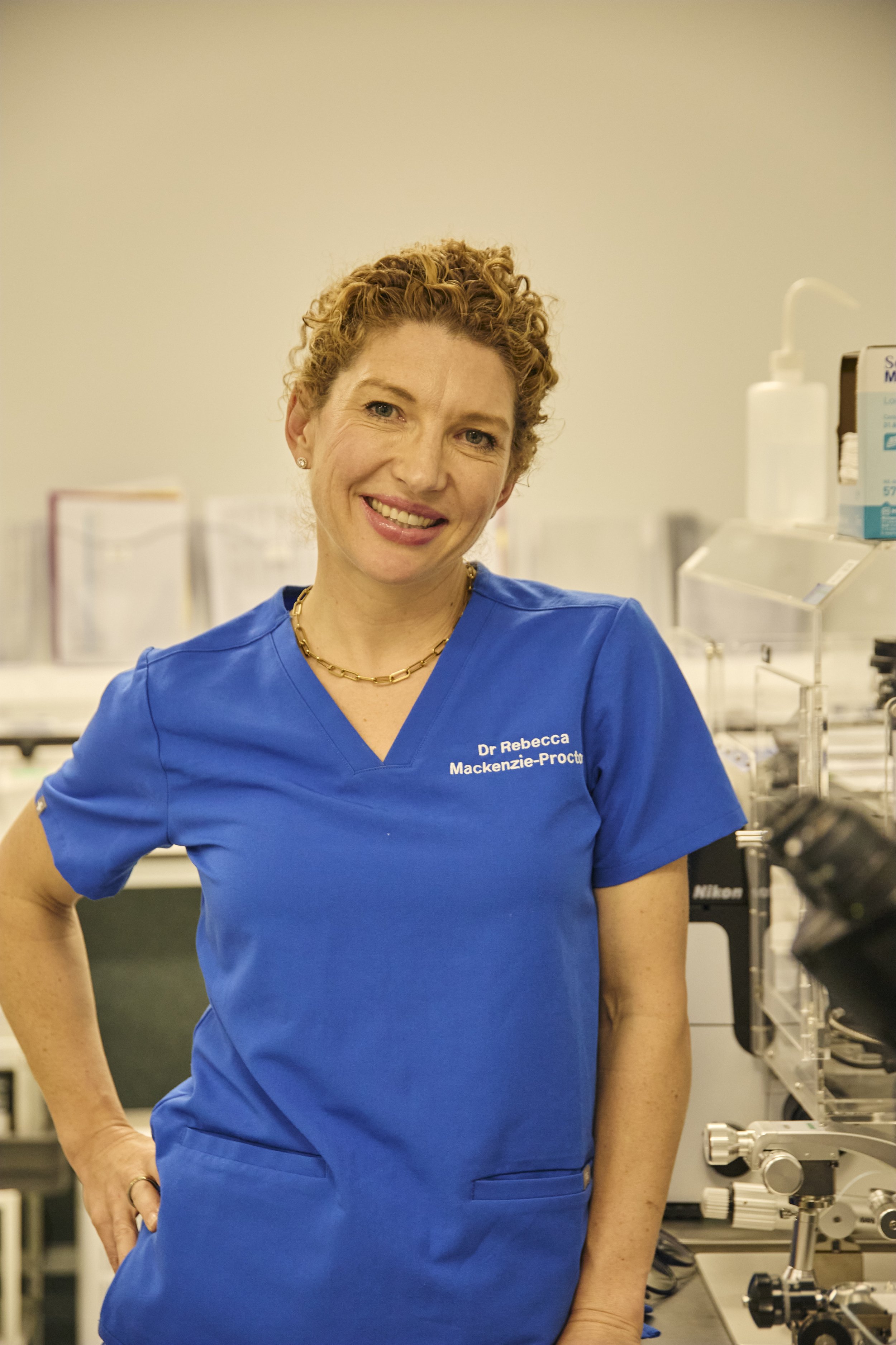 A woman wearing blue medical scrubs with the name "Dr. Rebecca Mackenzie-Proctor" embroidered on it, standing in a laboratory or medical setting.
