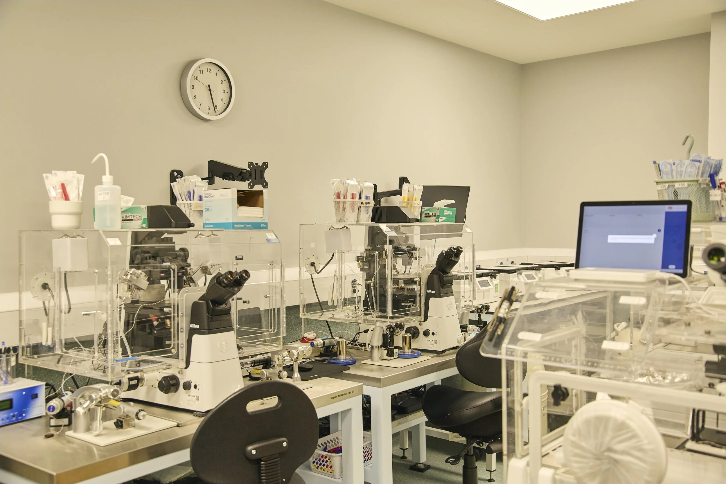 Laboratory with microscopes, equipment, and supplies on white tables, a wall clock, and a computer monitor on the right side.