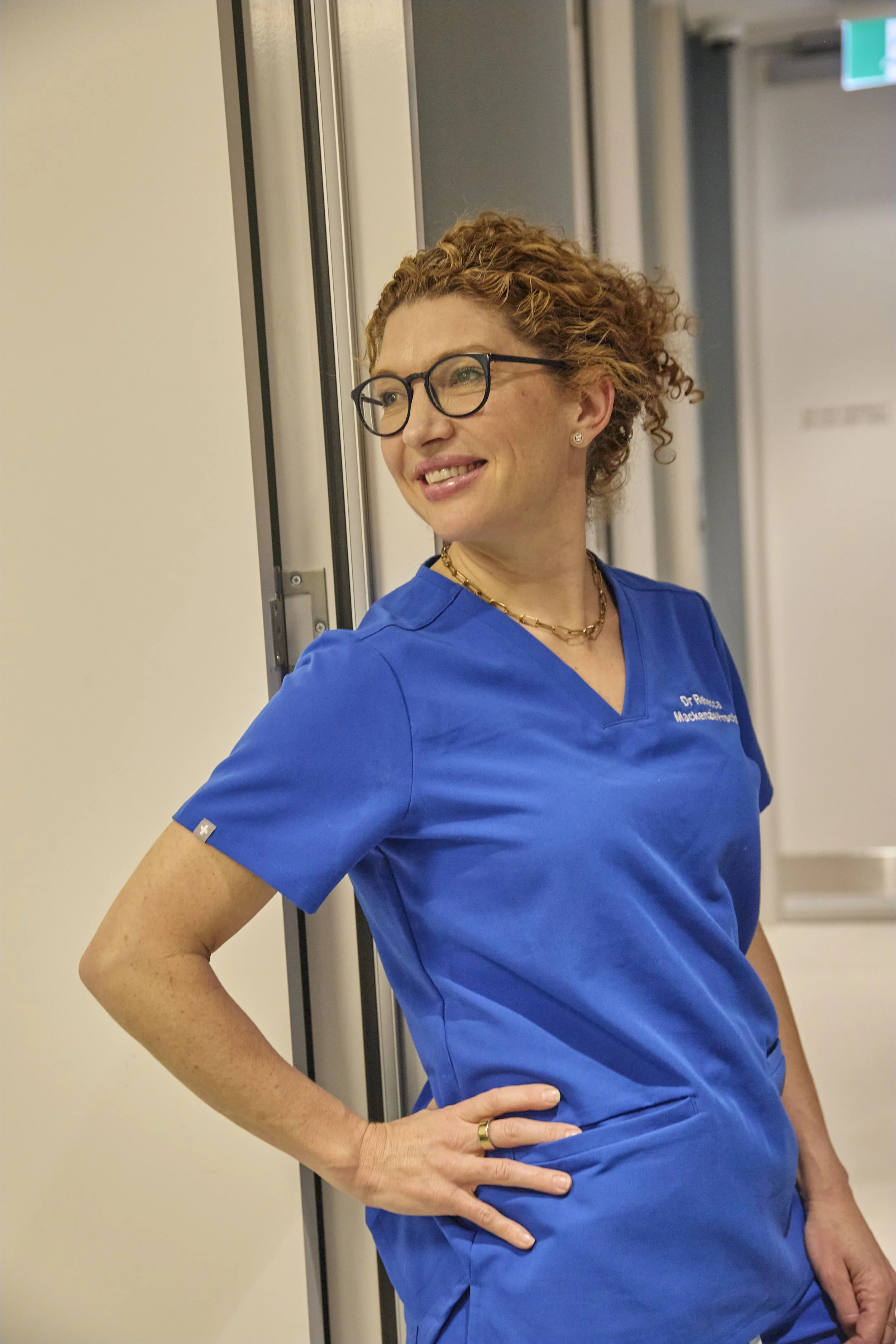 A woman with curly reddish hair, glasses, and wearing blue medical scrubs stands in a hospital or clinic corridor, smiling and looking off to the side.
