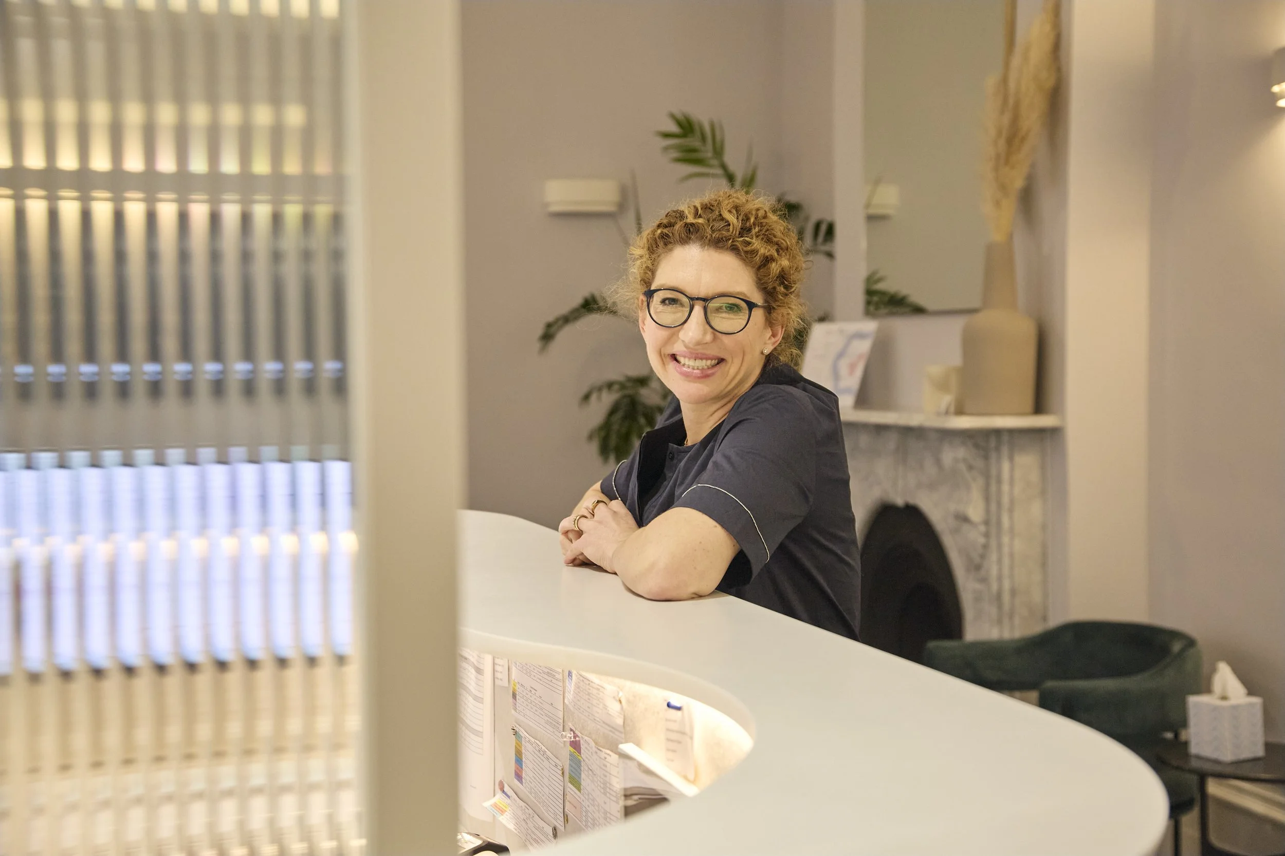 A smiling woman with curly red hair, glasses, and a black shirt sitting at a curved white reception desk in a warmly lit room with decorative plants and a fireplace in the background.