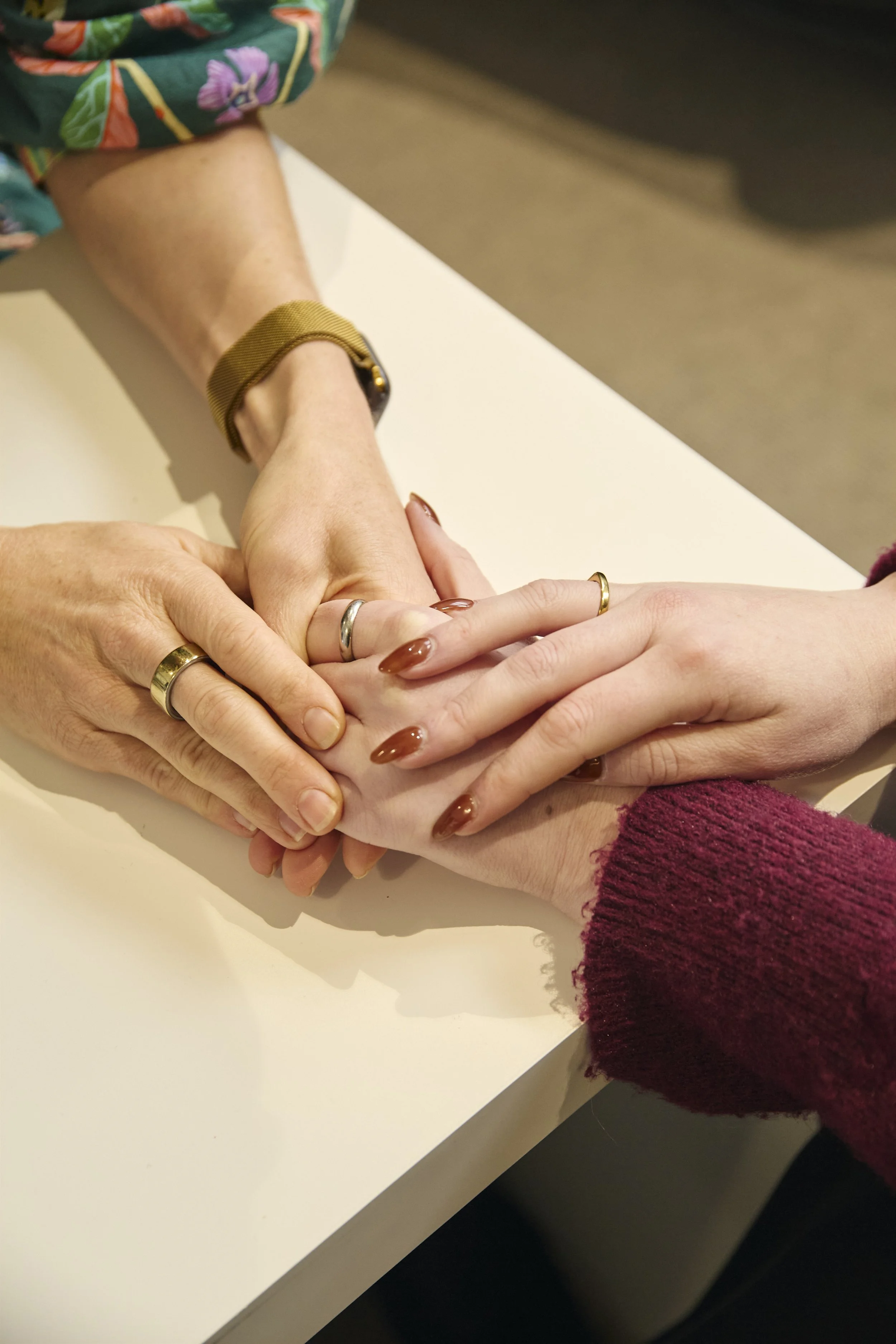 Three people with intertwined hands, wearing rings, on a white table, one person with a brown jacket and long, manicured nails, another with a maroon sweater and a gold bangle.