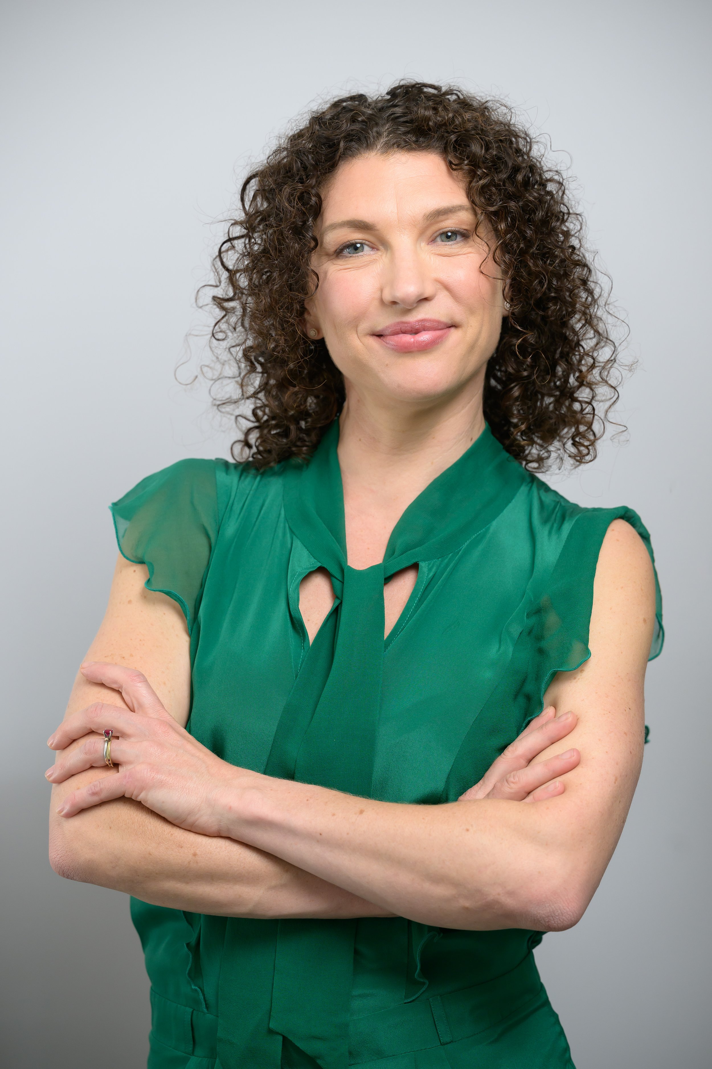 A woman with curly brown hair, smiling, wearing a sleeveless green blouse with ruffled cap sleeves and a matching necktie, standing against a light gray background, with arms crossed.