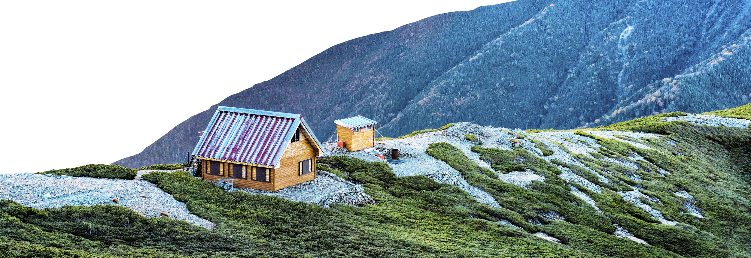 A small wooden cabin with a metal roof and a smaller shed on a hillside covered in green bushes and rocks, with mountainous terrain in the background.