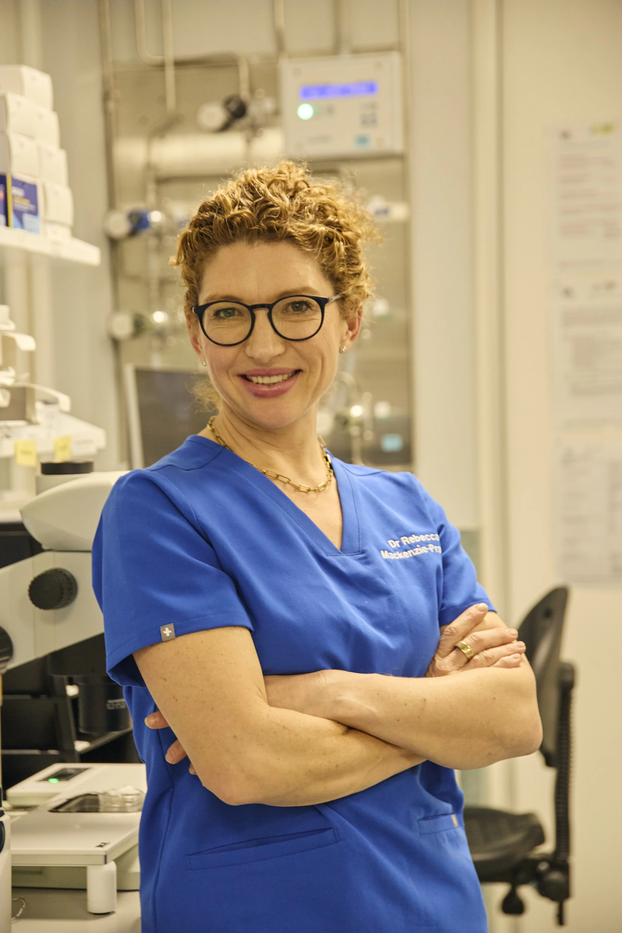 A female healthcare professional with short curly red hair and glasses, wearing a blue medical scrub top, standing with arms crossed in a hospital or medical facility.