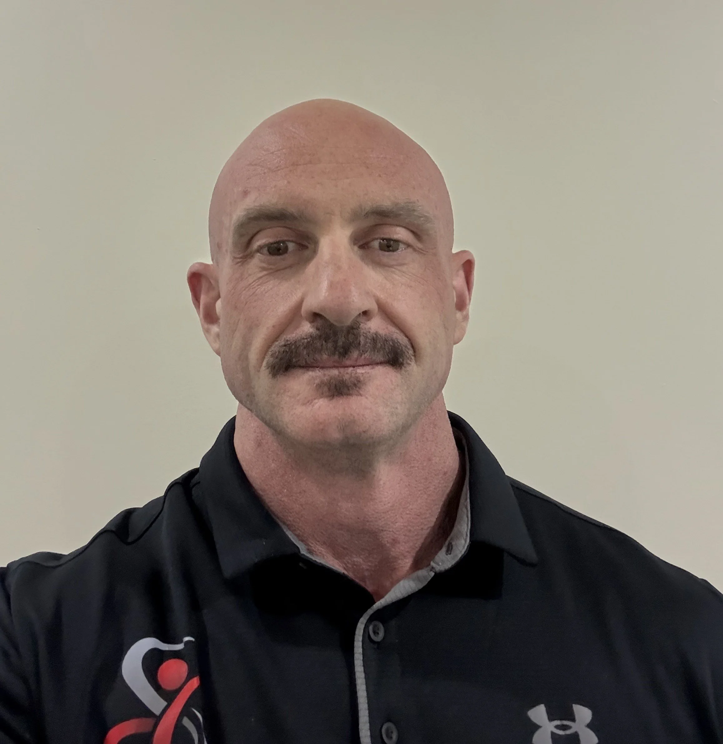 A bald man with a mustache and goatee wearing a black collared shirt with a logo on the chest is taking a selfie against a plain light-colored background.