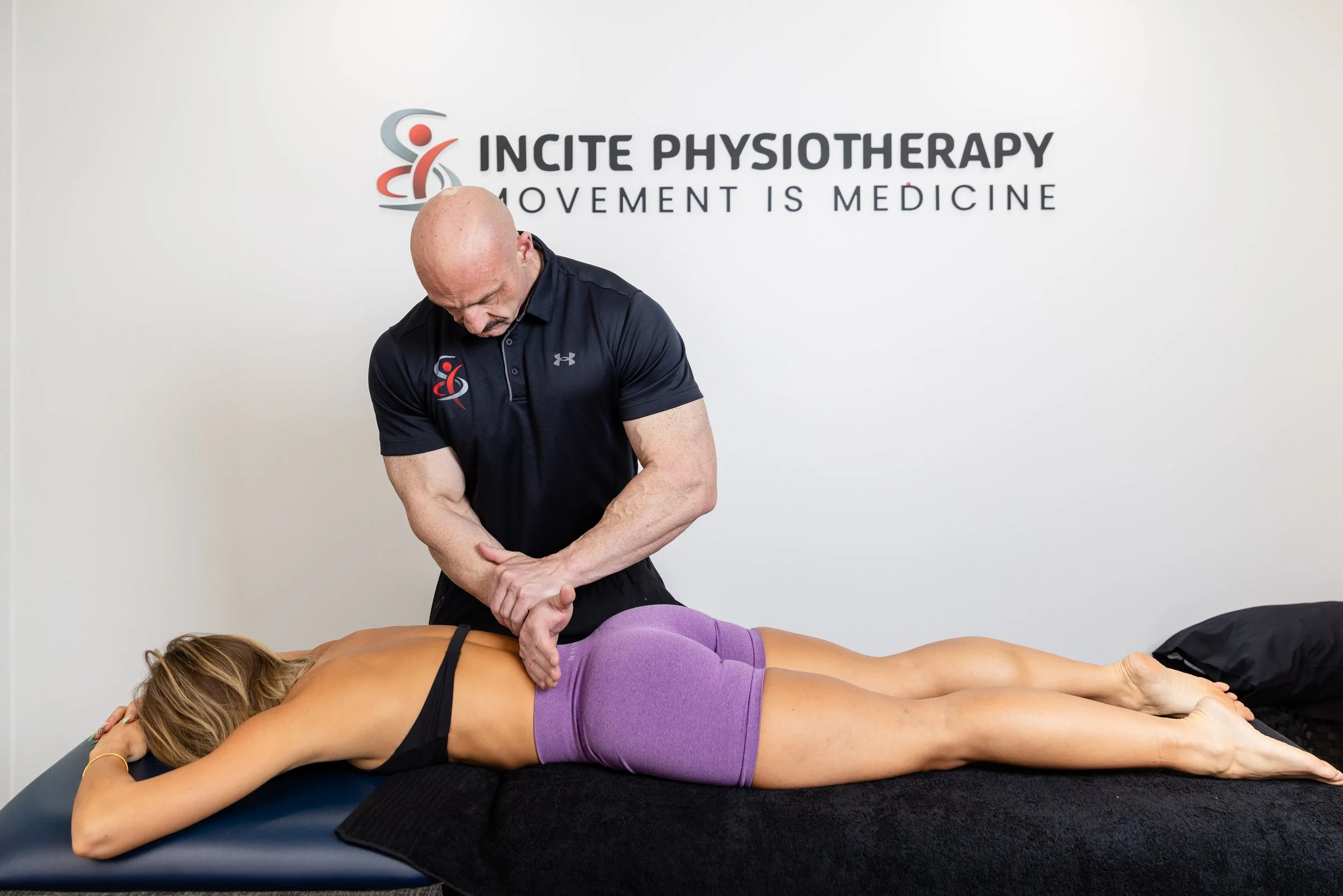 A male physiotherapist is performing treatment on a woman lying face down on a black treatment table, with her head resting on her arm, in a clinic with a sign that reads 'Incite Physiotherapy, Movement is Medicine.'