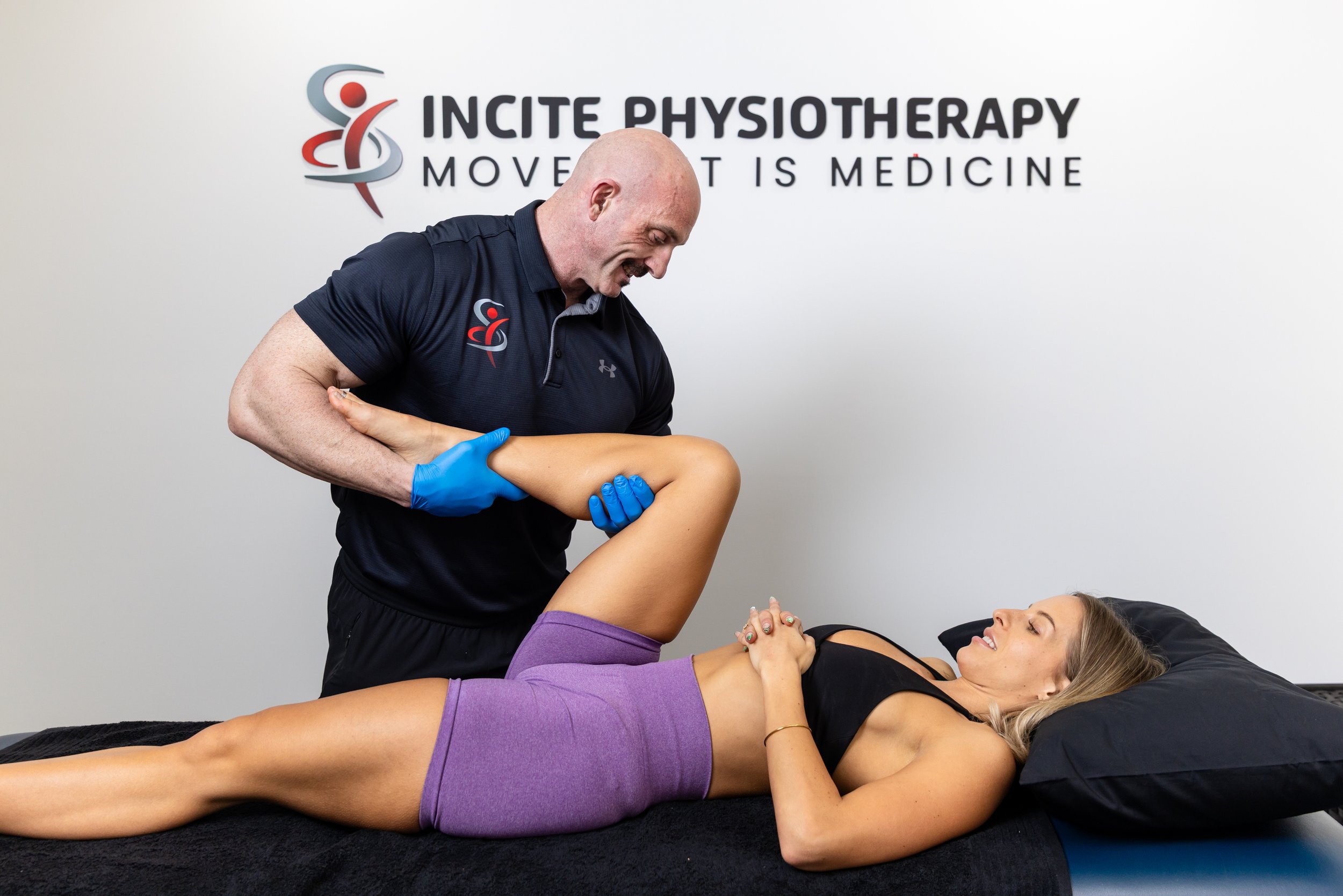 A physiotherapist in a black shirt with blue gloves lifting a woman's leg during a physiotherapy session in a clinic with a sign that says 'Incite Physiotherapy.'