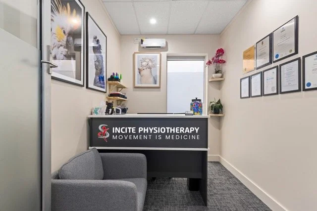 Reception area with a gray armchair, framed artwork on the walls, and a sign that reads 'Incite Physiotherapy: Movement is Medicine' behind the reception desk.