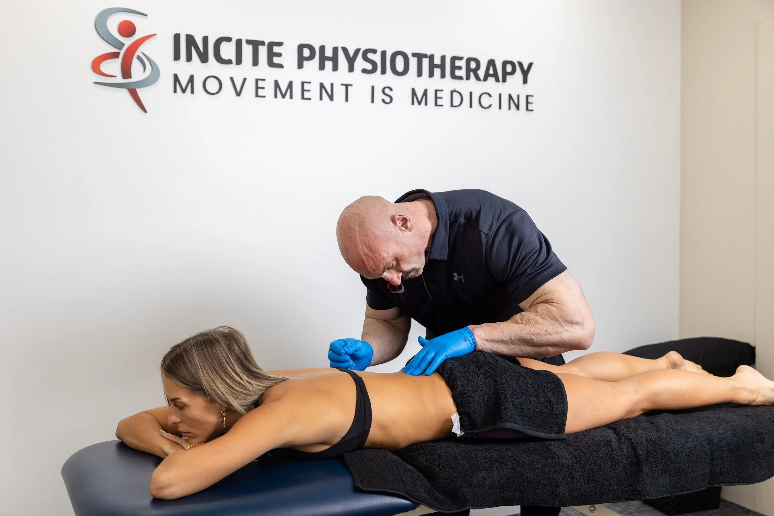 A physiotherapist in black shirt with blue gloves performs a treatment on a woman lying face down on a massage table, in a room with 'Incite Physiotherapy' sign on the wall.