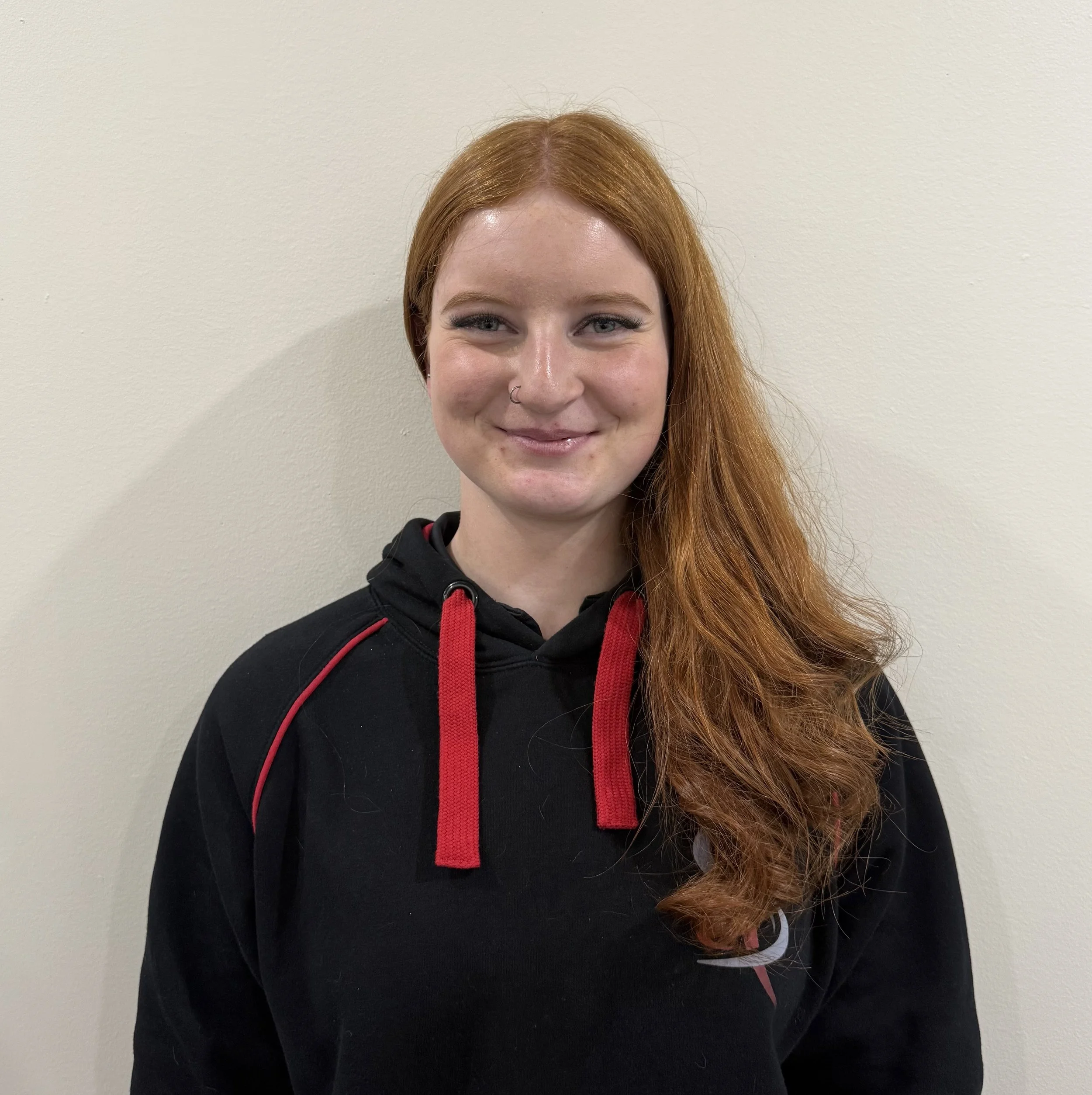 A young woman with long, wavy red hair, fair skin, and a nose piercing, smiling and wearing a black hoodie with red drawstrings, standing against a plain beige wall.