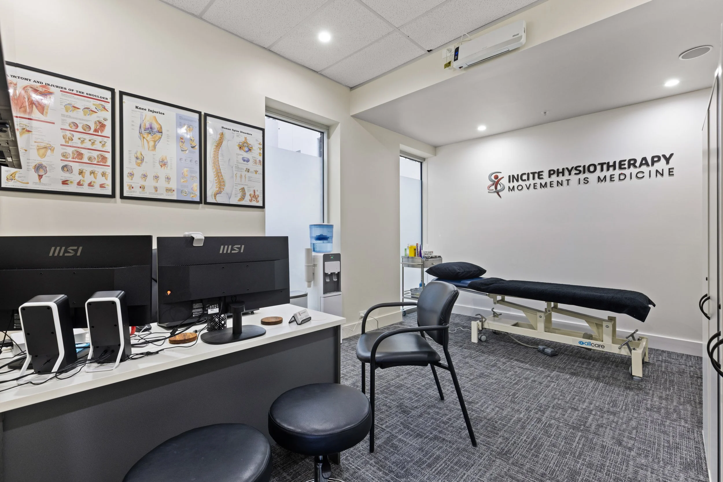 Interior of a physiotherapy clinic with therapy table, two black chairs, computer monitors, framed anatomy posters on the wall, water dispenser, and a sign on the wall reading 'Incite Physiotherapy Movement is Medicine'.