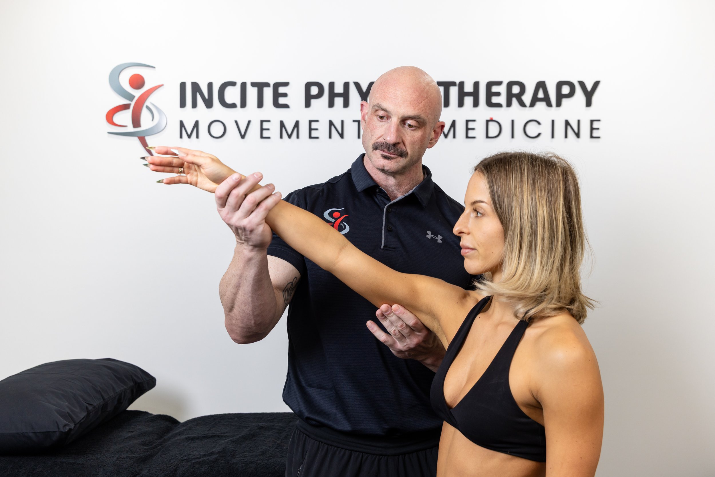 A man assisting a woman with shoulder stretch exercises in a physiotherapy clinic, with 'Incite Physiotherapy Movement Medicine' sign in the background.