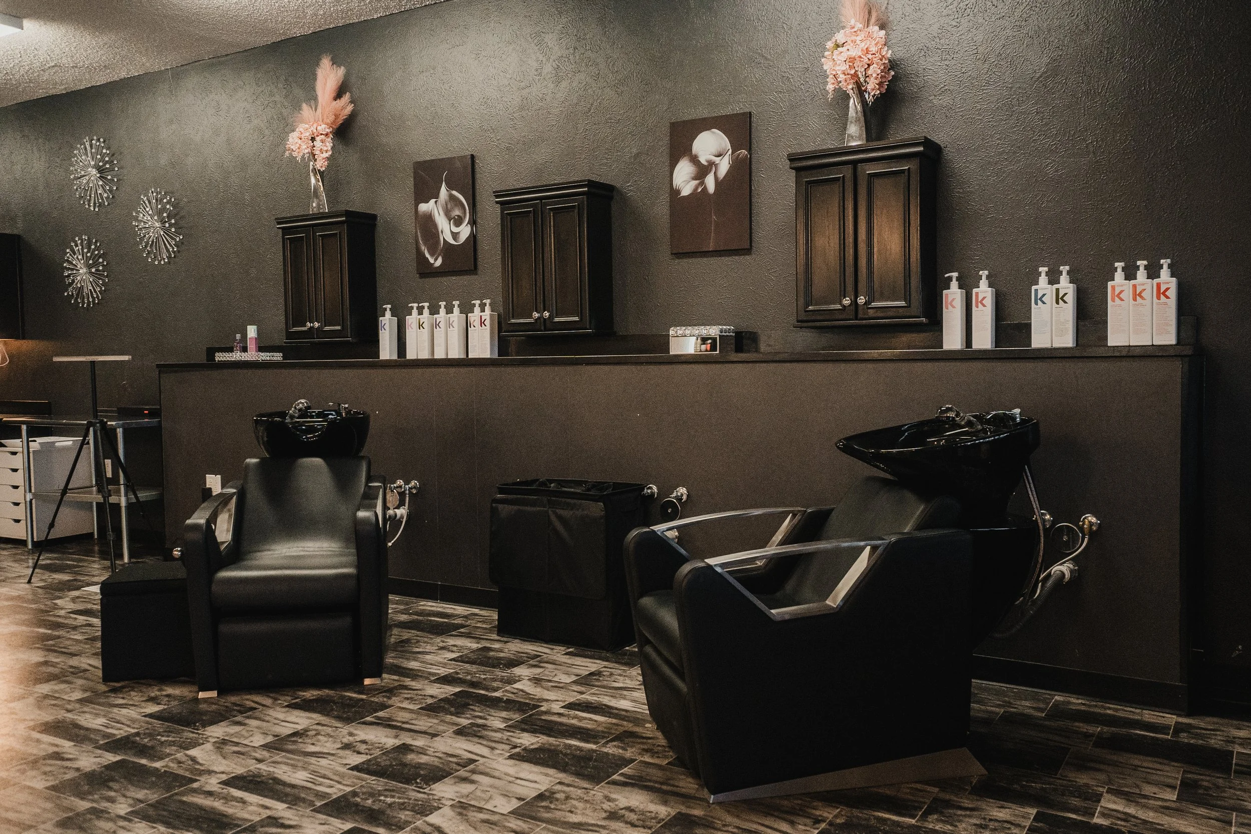 Interior of a hair salon with black reclining chairs and washing basins, black cabinets, wall decorations, and bottles of hair care products.