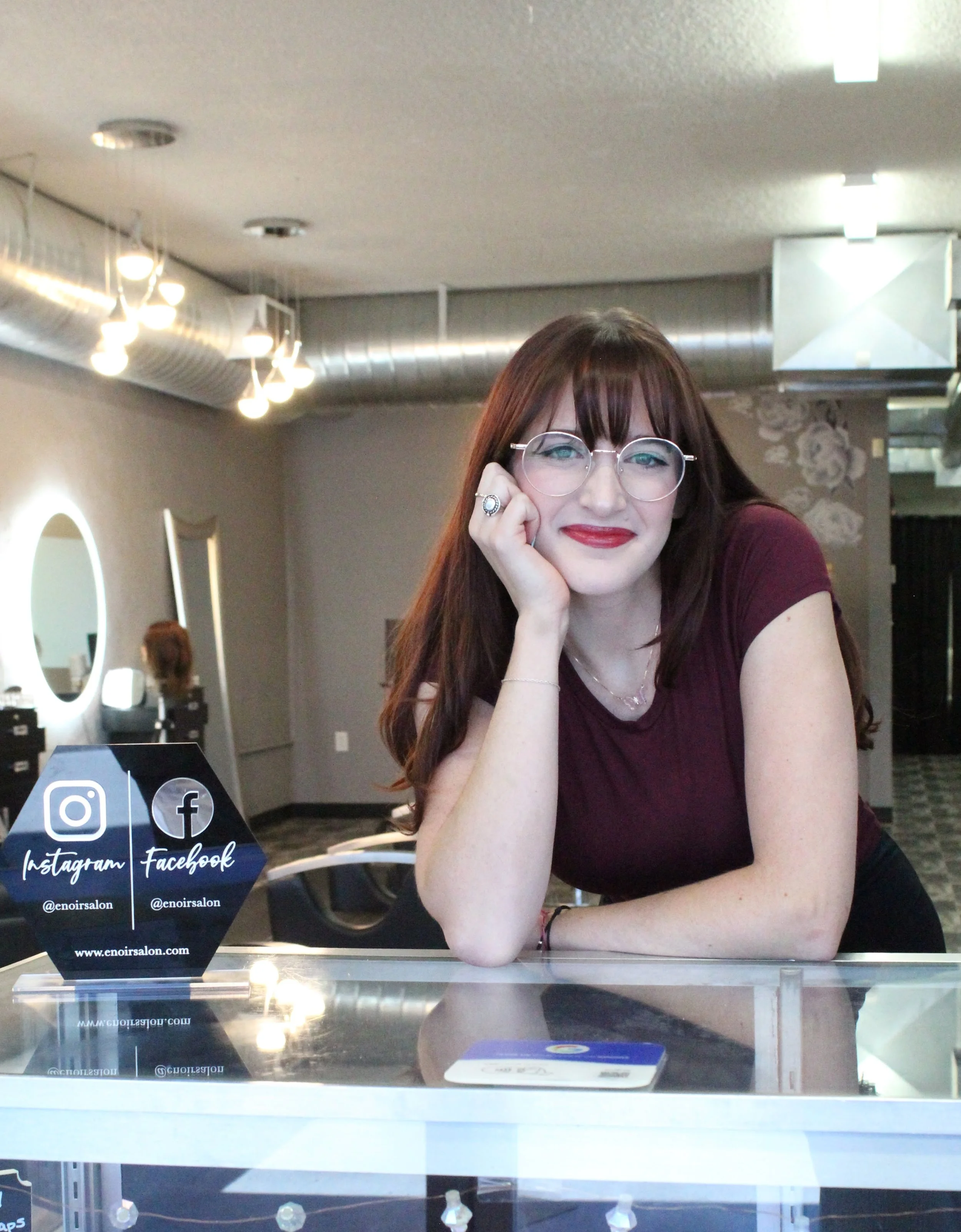 Katie,  with long reddish-brown hair, glasses, and a burgundy top sitting at a front desk in a salon. She is smiling and resting her head on her hand. The salon has modern decor with round mirrors, hanging light fixtures, and ventilation ducts.