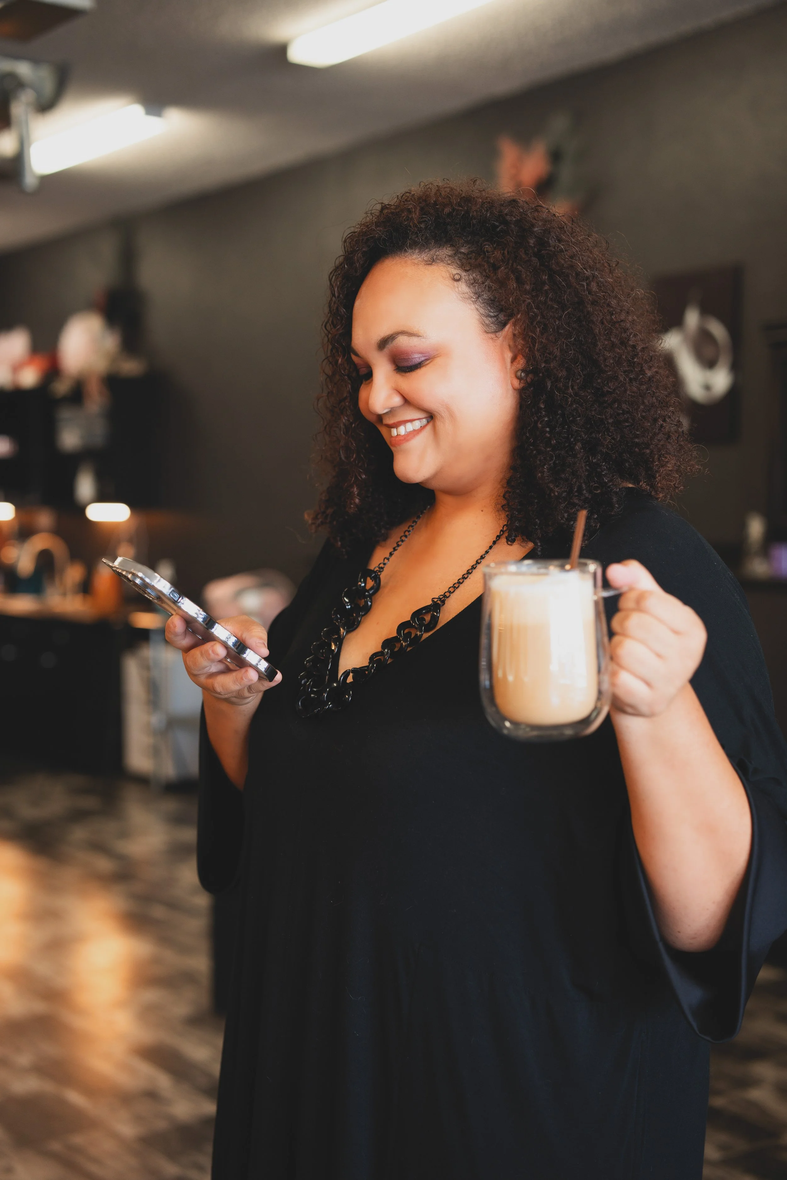 A woman with curly hair smiling while looking at her phone, holding a glass mug of coffee or latte in her other hand, inside a coffee shop or cafe.