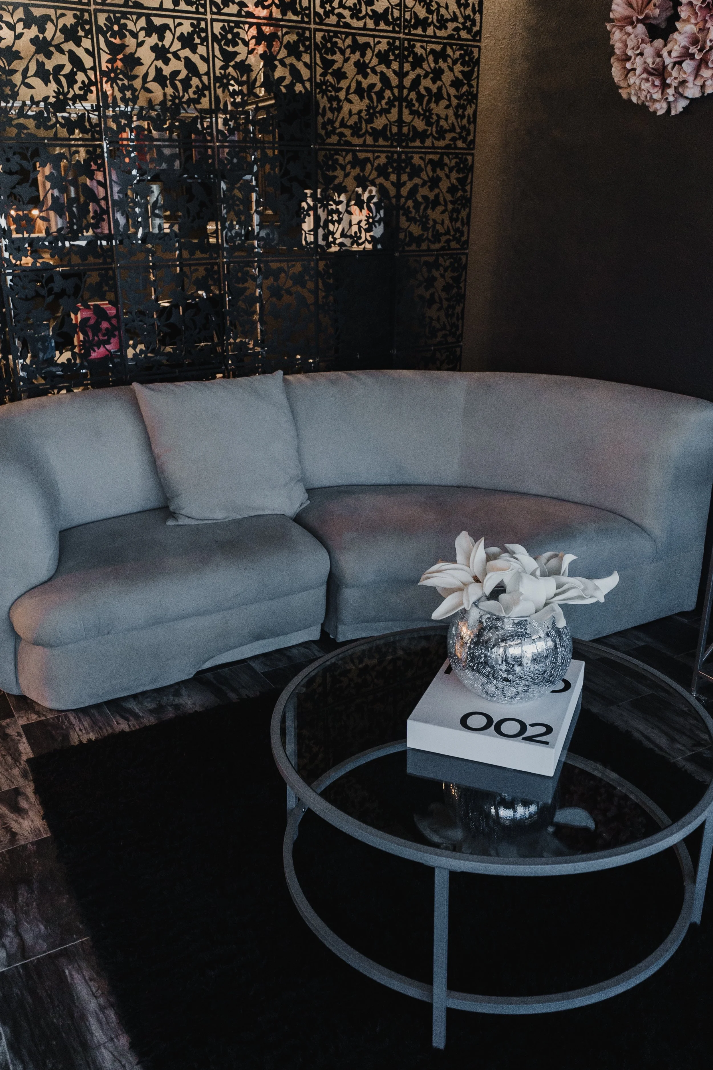 A cozy corner with a light gray sectional sofa with a matching pillow, a round glass coffee table with a white book and a silver vase with white lilies, a black rug, and a decorative black partition with floral cutouts in the background.