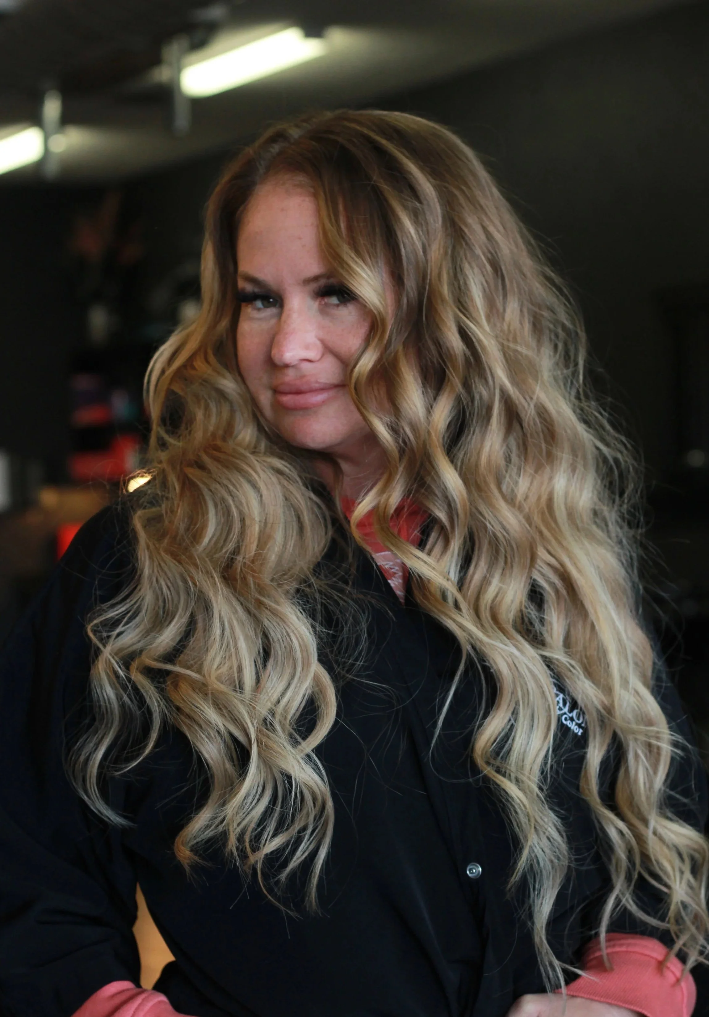 A woman with long, wavy blonde hair extensions and freckles looks at the camera in a dimly lit indoor setting.