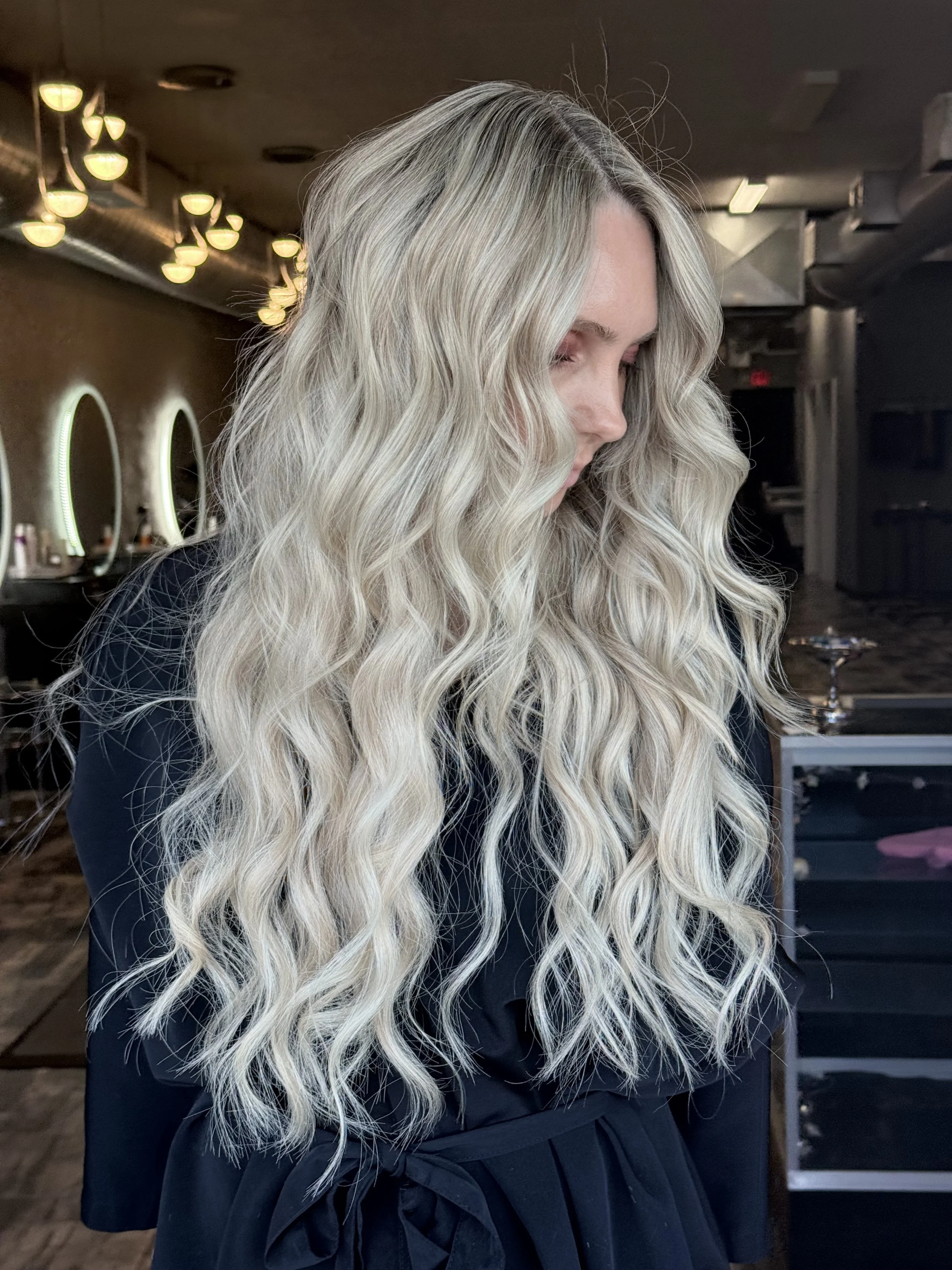 A woman with long, wavy platinum blonde hair looking down, wearing a black outfit, in a salon with circular mirrors and warm lighting.