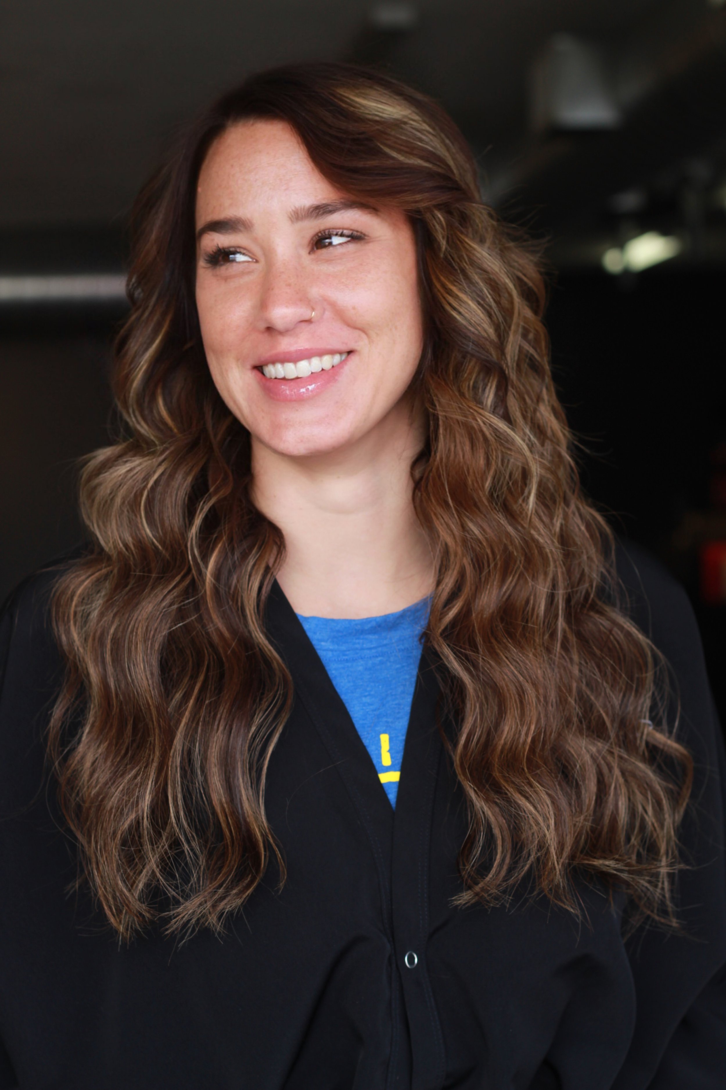 A woman with long wavy brown hair, wearing a blue shirt and a black jacket, smiling and looking to the side.