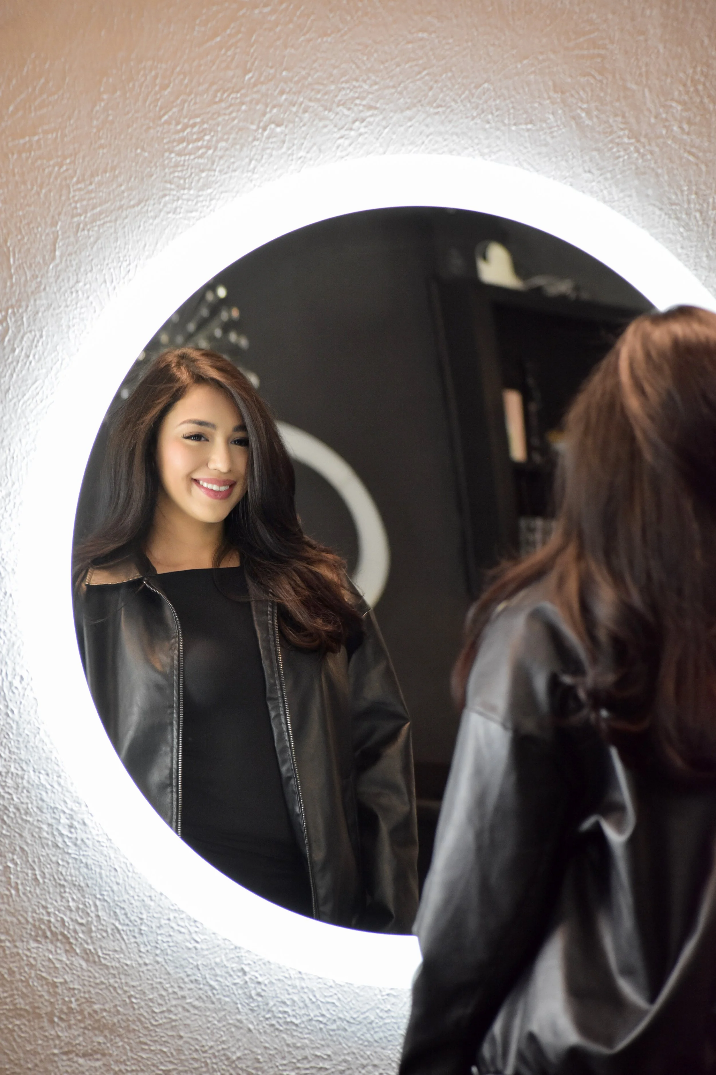 Jiseell, stylist with long brown hair smiling at her reflection in a round mirror, in a room with textured white wall.