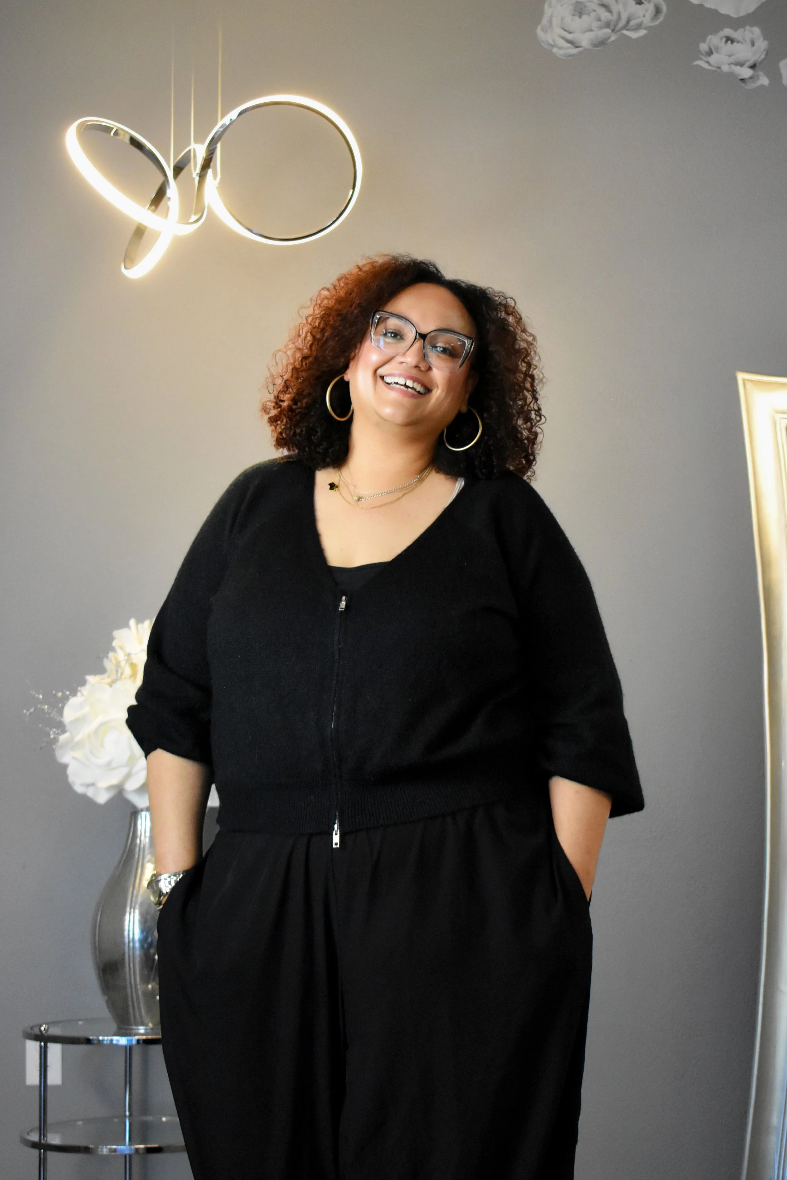 Ginger, with curly hair, glasses, and hoop earrings smiling at the camera, standing in front of a gray wall with a modern circular ceiling light and a gold-framed mirror.