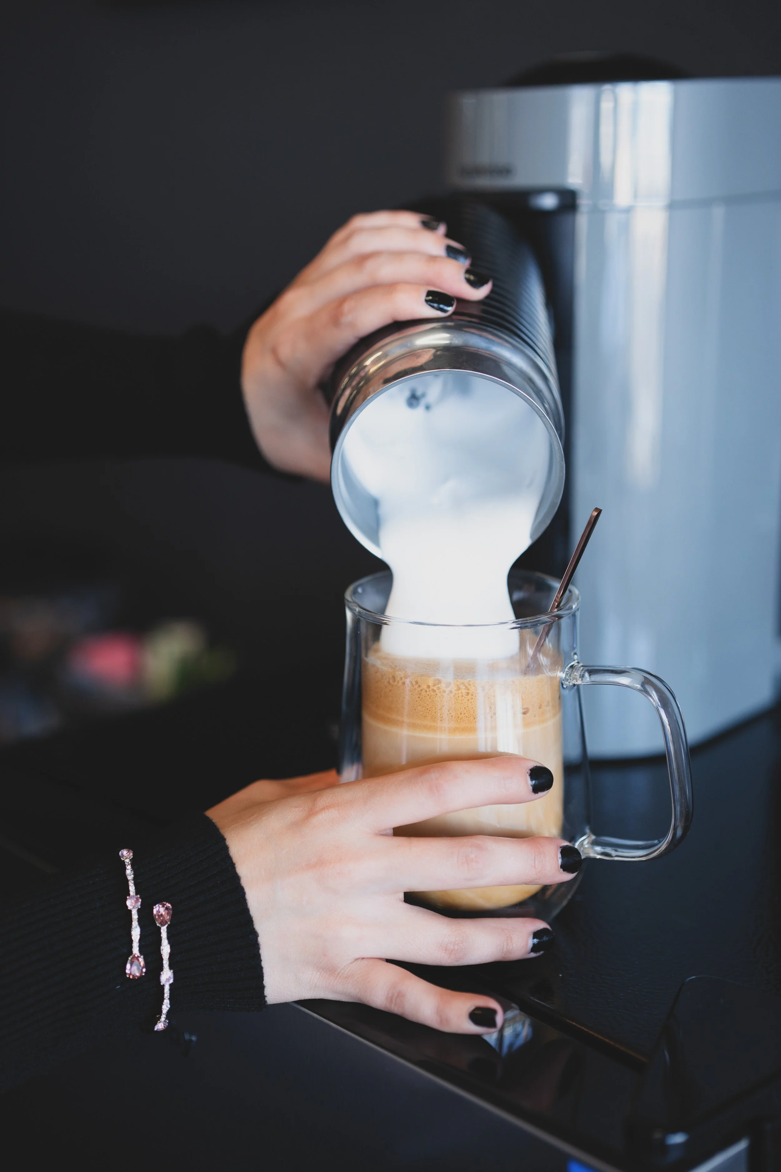 A person with black painted nails is pouring milk into a glass mug of coffee using a metal pitcher. The person is wearing a black sleeve with a pink gemstone bracelet.