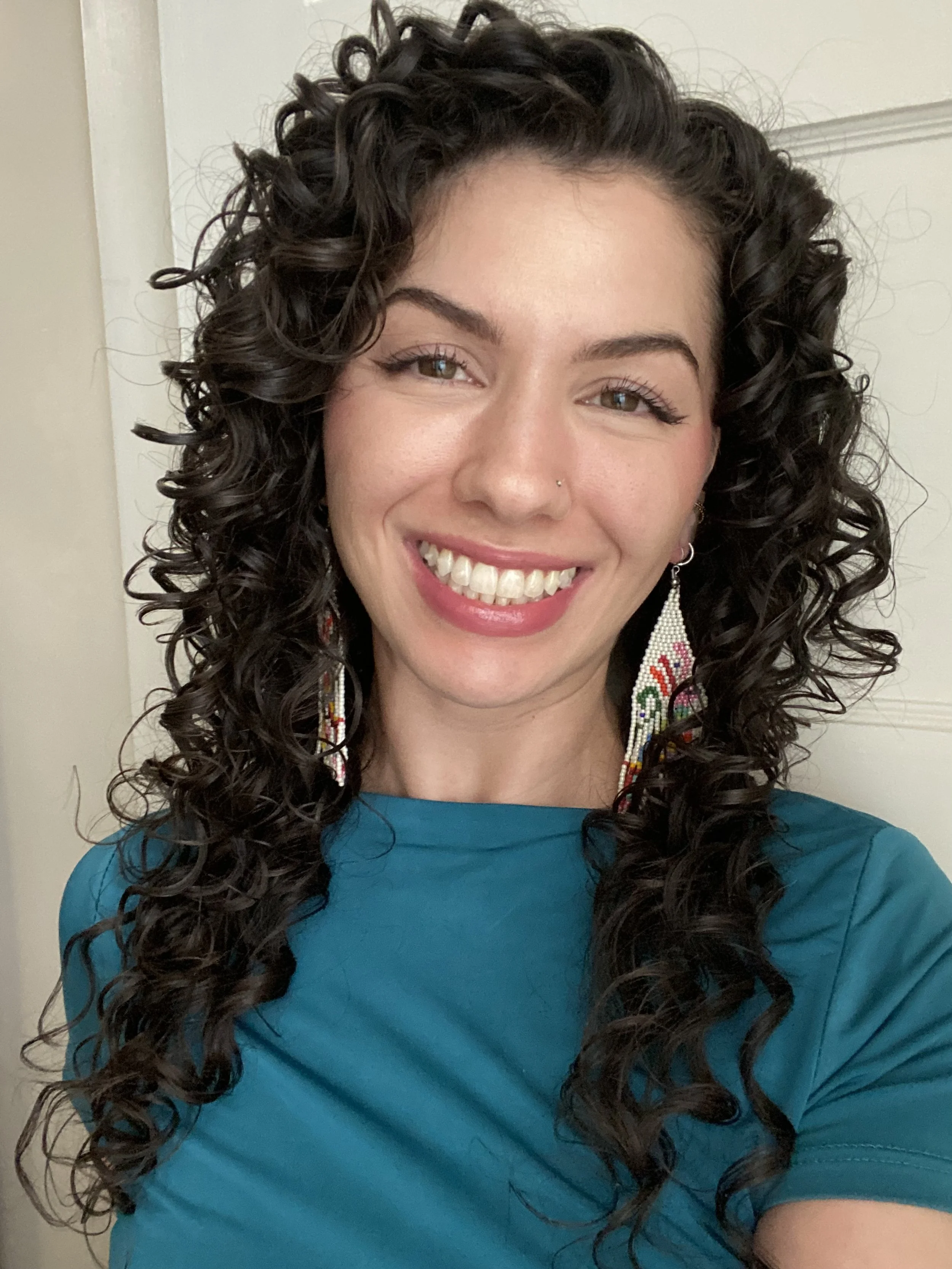 A young woman with long, dark, curly hair smiling at the camera, wearing a teal shirt and colorful beaded earrings.