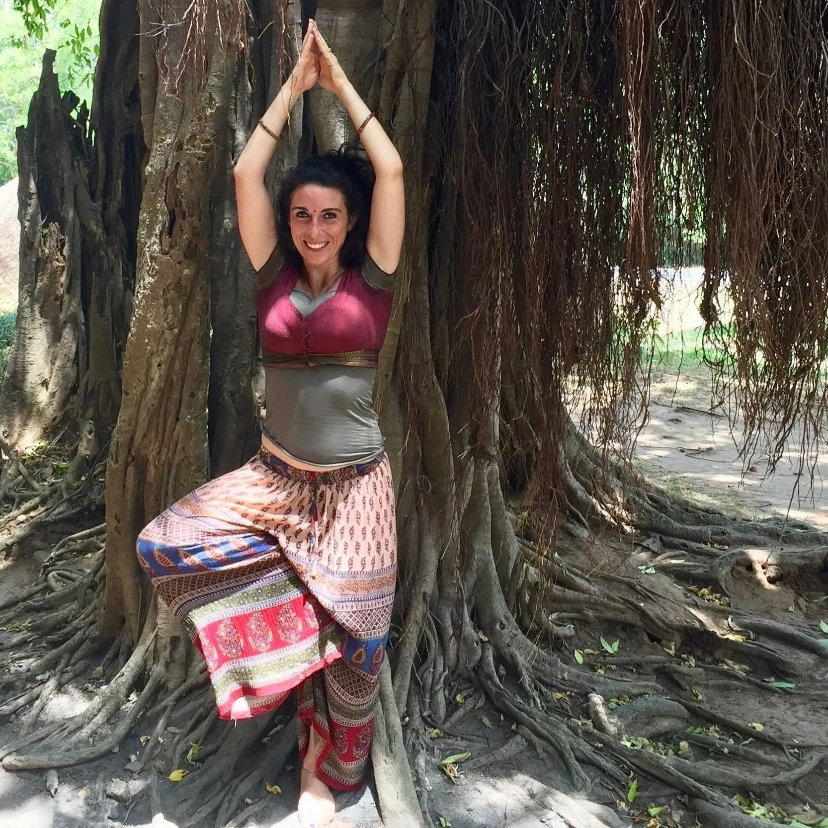 Una donna sorridente, con capelli neri lunghi e vestiti colorati, si trova vicino a un grande albero con radici visibili, in un ambiente naturale con foglie verdi sullo sfondo.