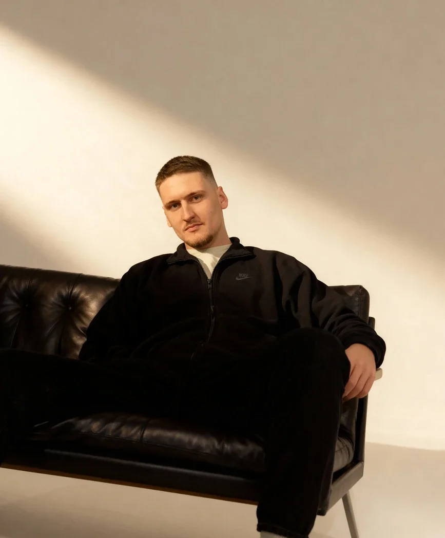 A young man with short hair and a goatee sitting on a black leather sofa against a plain light-colored wall, wearing a black Nike zip-up jacket and black pants.