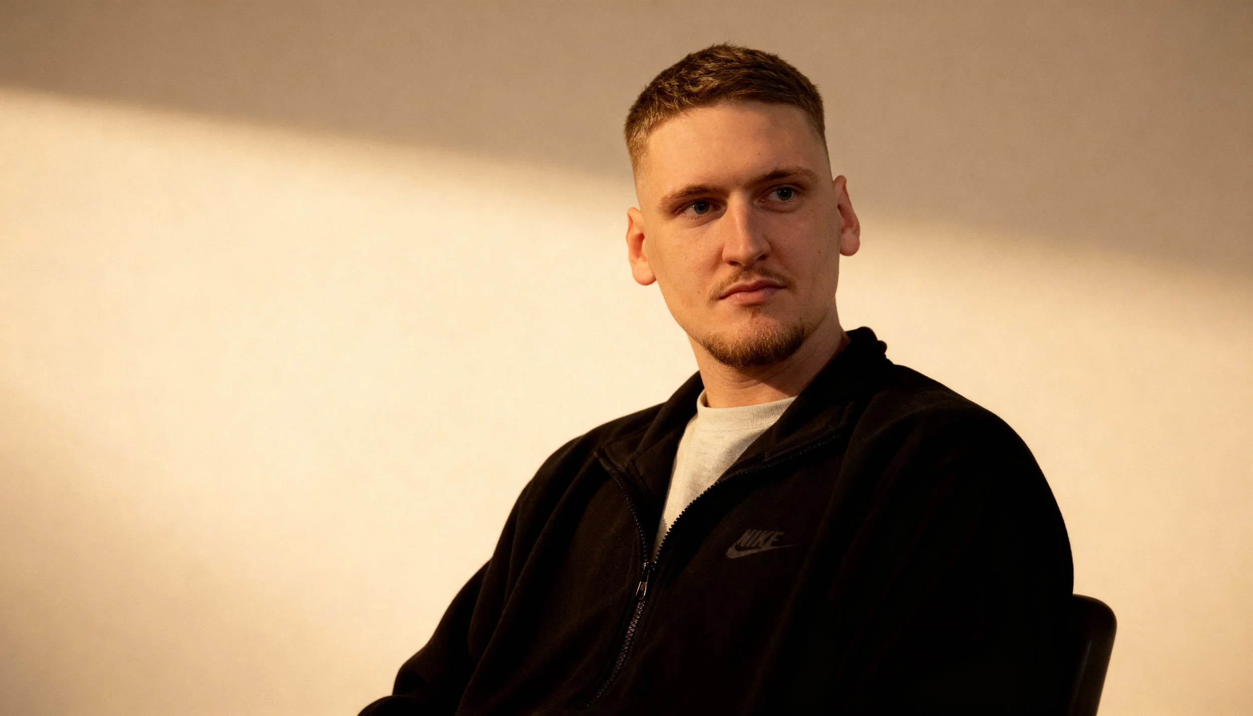 A young man with short hair and a goatee, wearing a black Nike zip-up jacket over a light-colored T-shirt, sitting against a plain background with a soft shadow.
