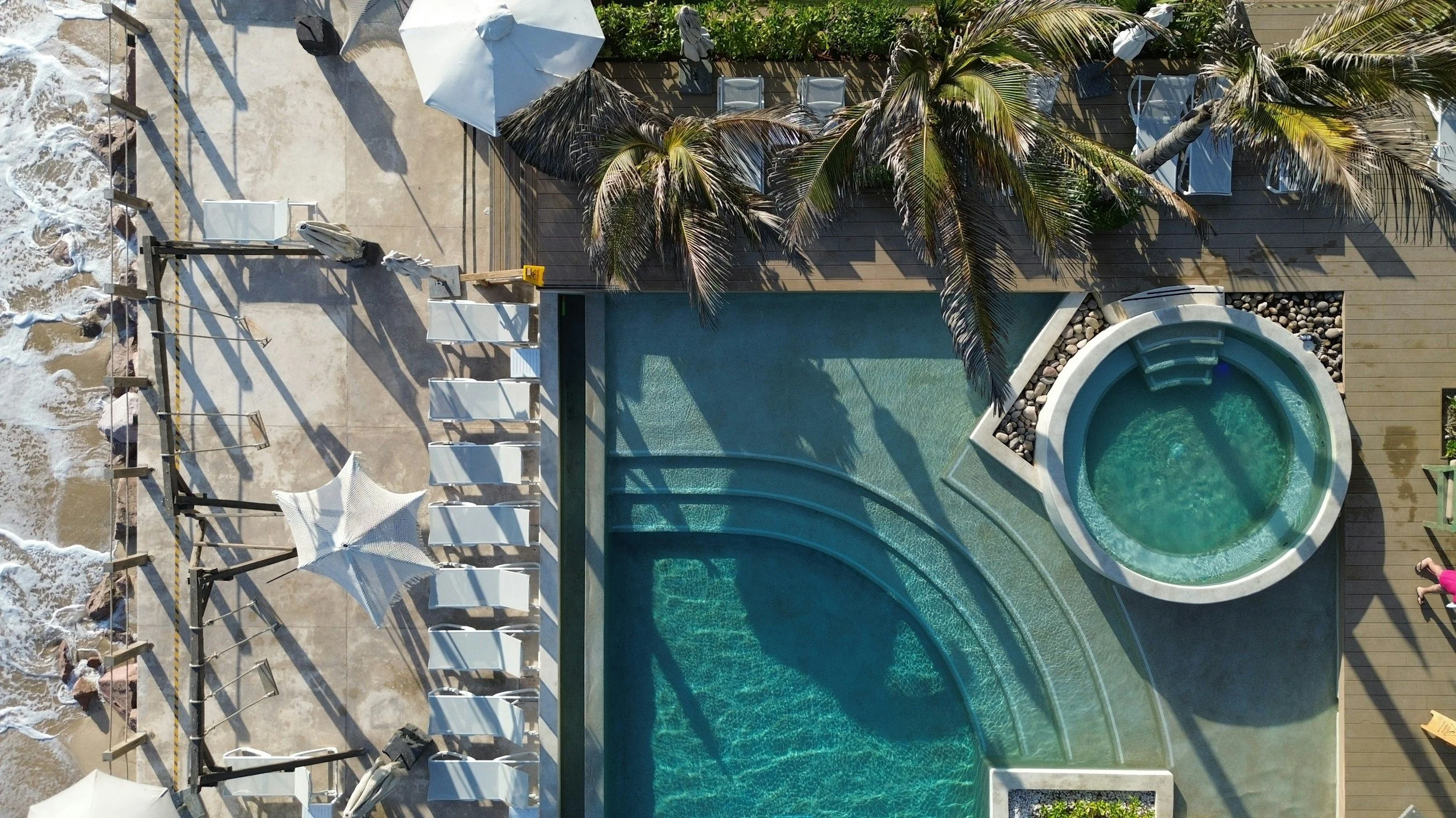Aerial view of a tropical outdoor pool area with a whirlpool, lounge chairs, umbrellas, and palm trees, adjacent to a sandy beach coastline with waves.