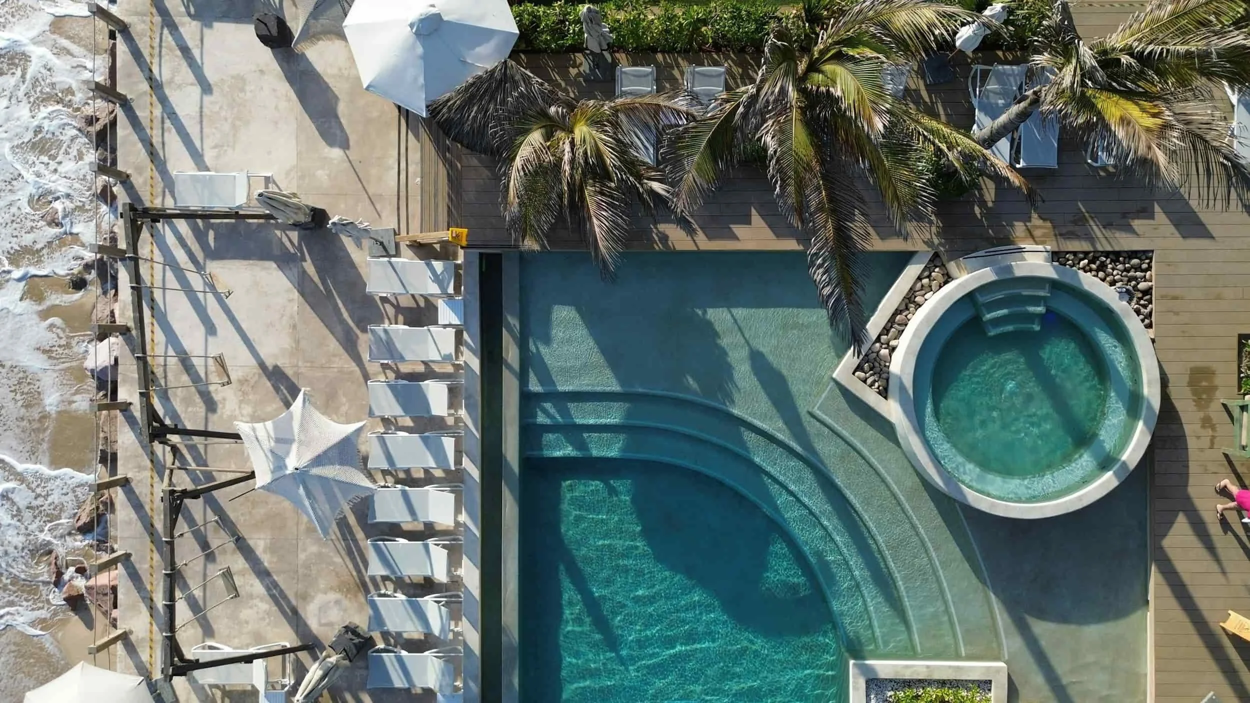 Aerial view of a pool and hot tub with palm trees, lounge chairs, umbrellas, and a Sunbed Booking System near the ocean.
