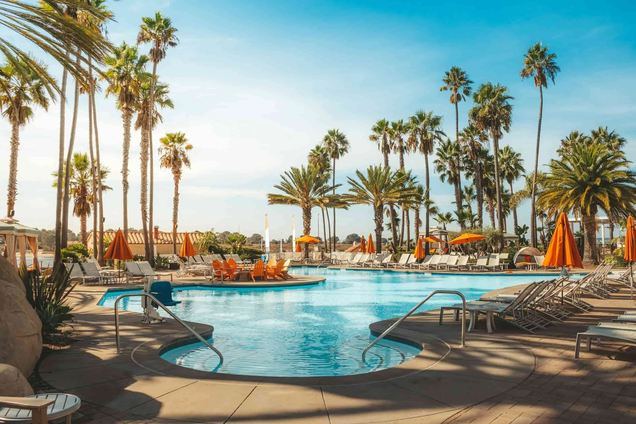 A resort swimming pool area with lounge chairs and orange umbrellas, tall palm trees, clear blue sky, and a swimming pool in the foreground.
