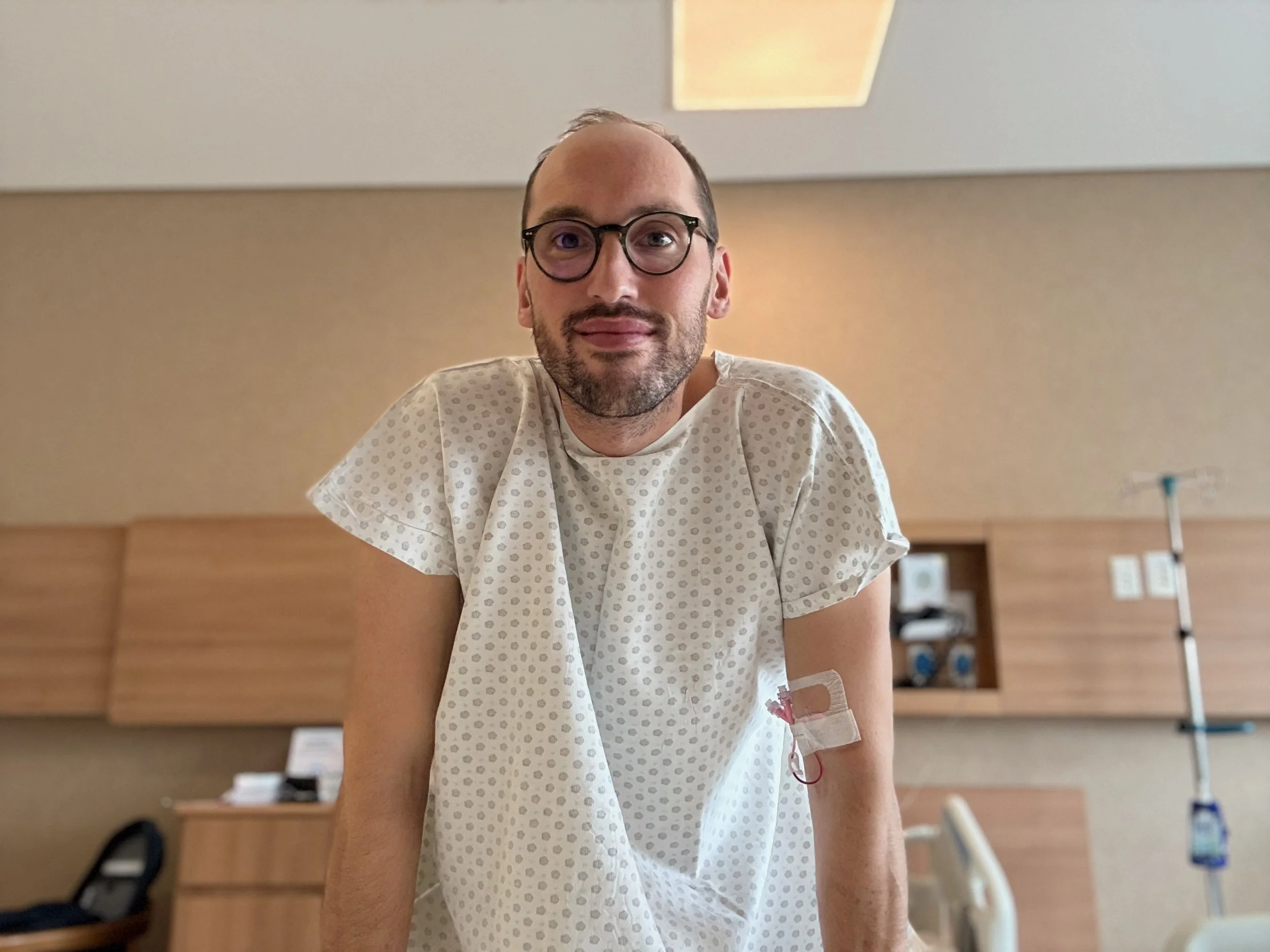 Ein Mann mit Brille und Bart liegt in einem Krankenhausbett, trägt einen Krankenhaushemd und hat eine Infusionsnadel in der Arm.