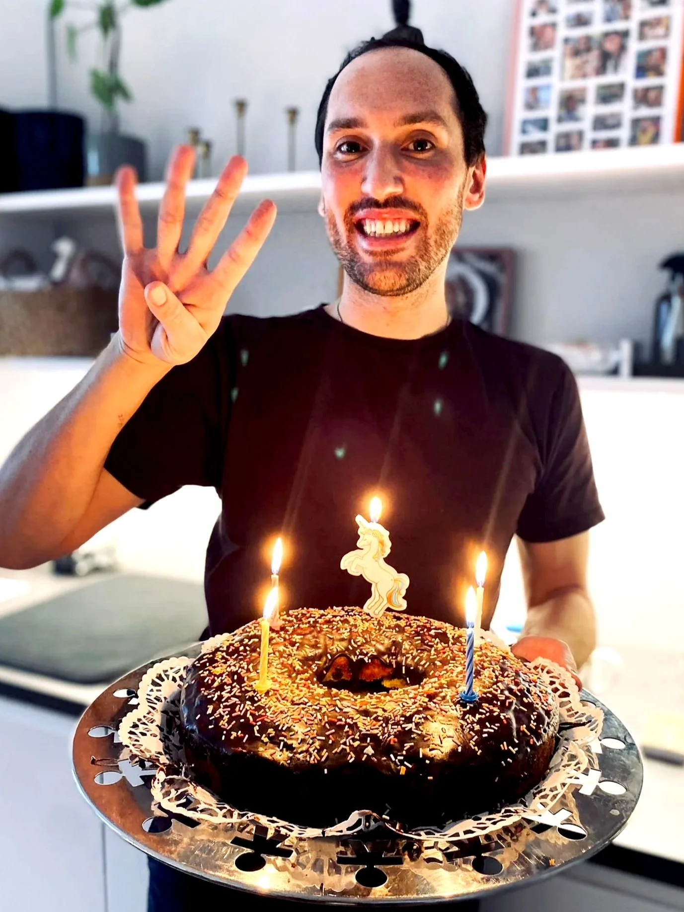 Junge Mann hält Geburtstagstorte mit Kerzen, auf der eine Einhorn-Feuerkerze brennt, und zeigt die vier Finger. Er lächelt und macht eine Victory-Geste. Im Hintergrund sind Küchenregale und Fotos an der Wand.