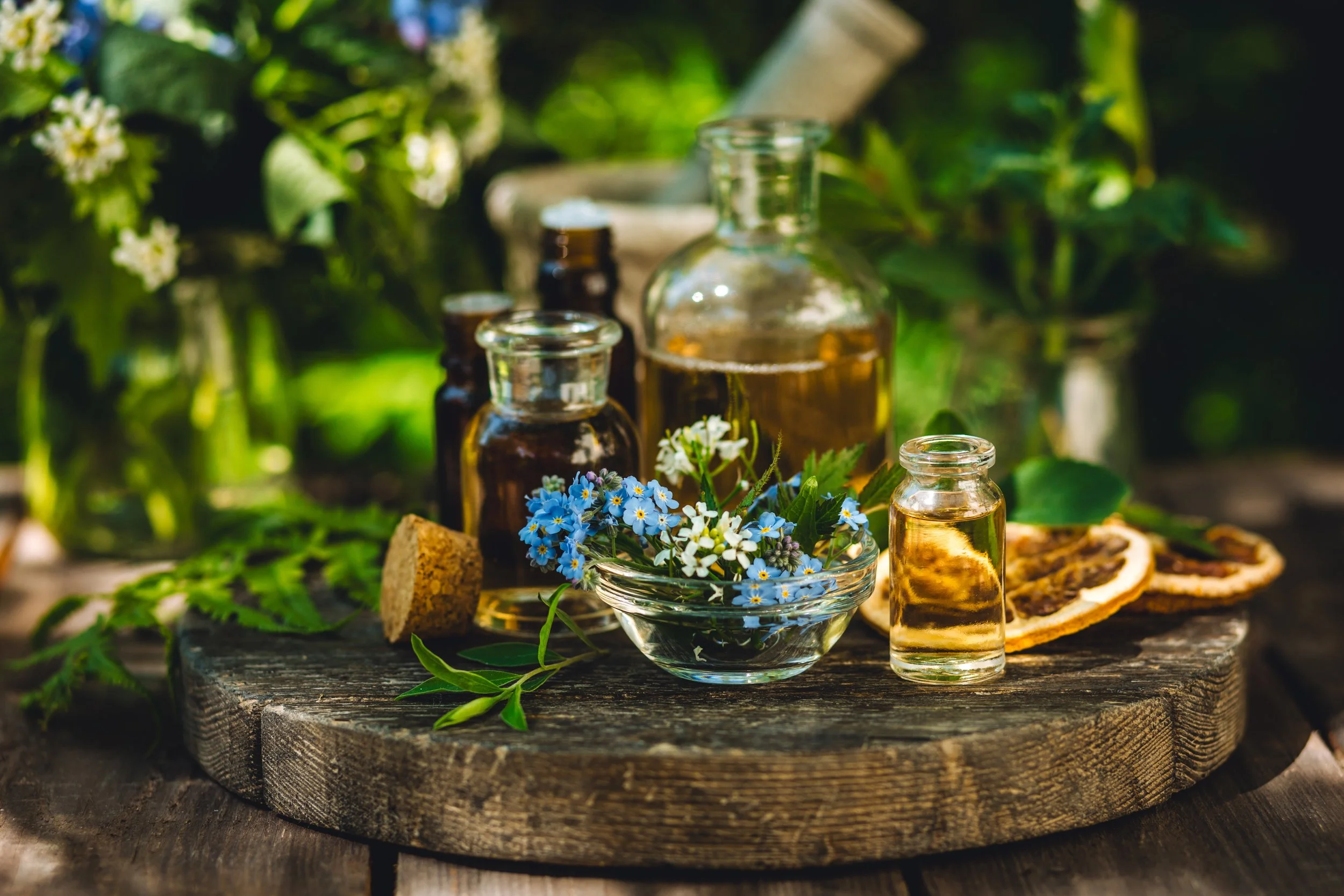 Assorted herbal and essential oils bottles with dried citrus slices, fresh herbs, and flowers on wooden surface outdoors.